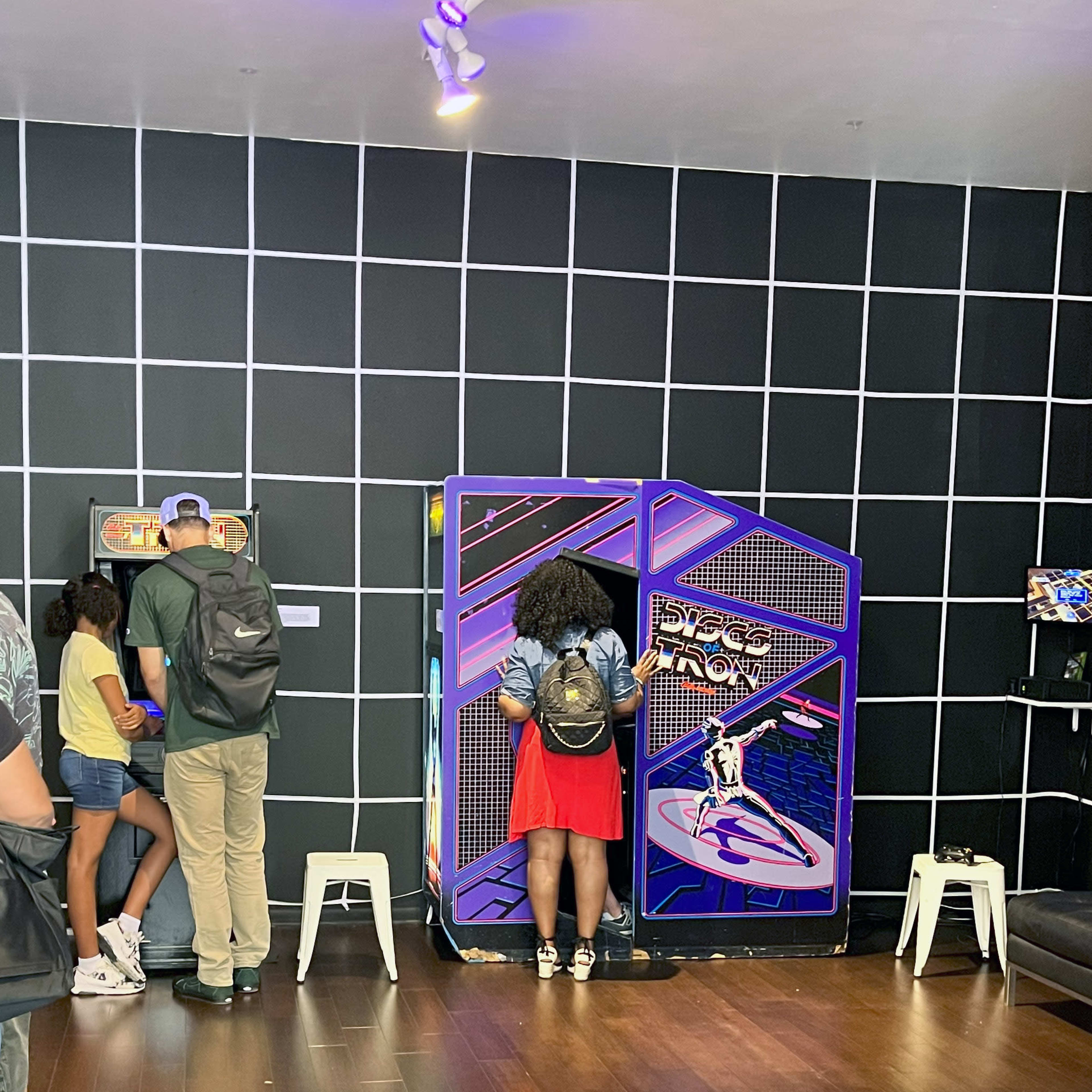 A group of people interacting with retro arcade games in a room with a grid-patterned black wall.