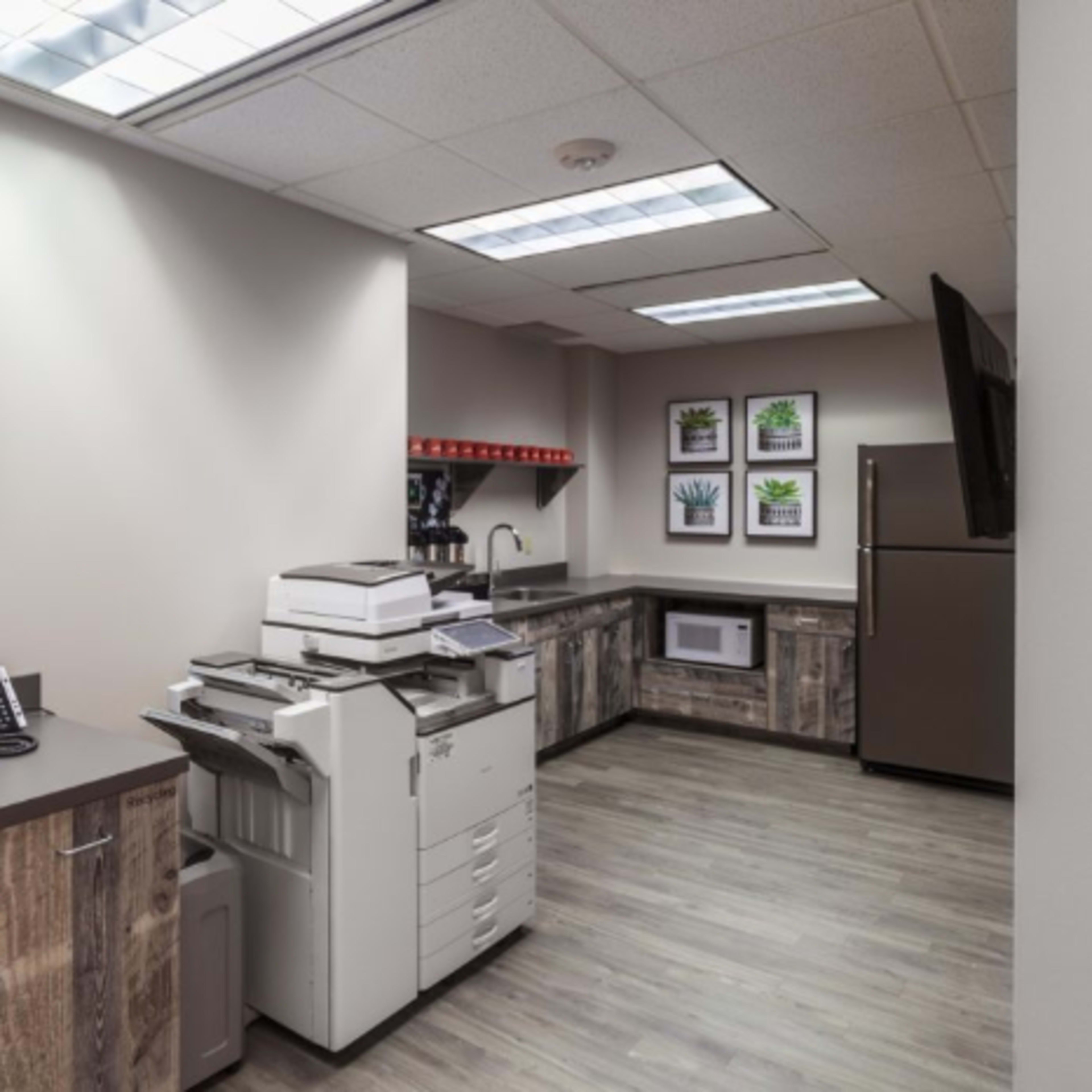An office break room with a copier, refrigerator, microwave, and a colorful dog painting on the wall.