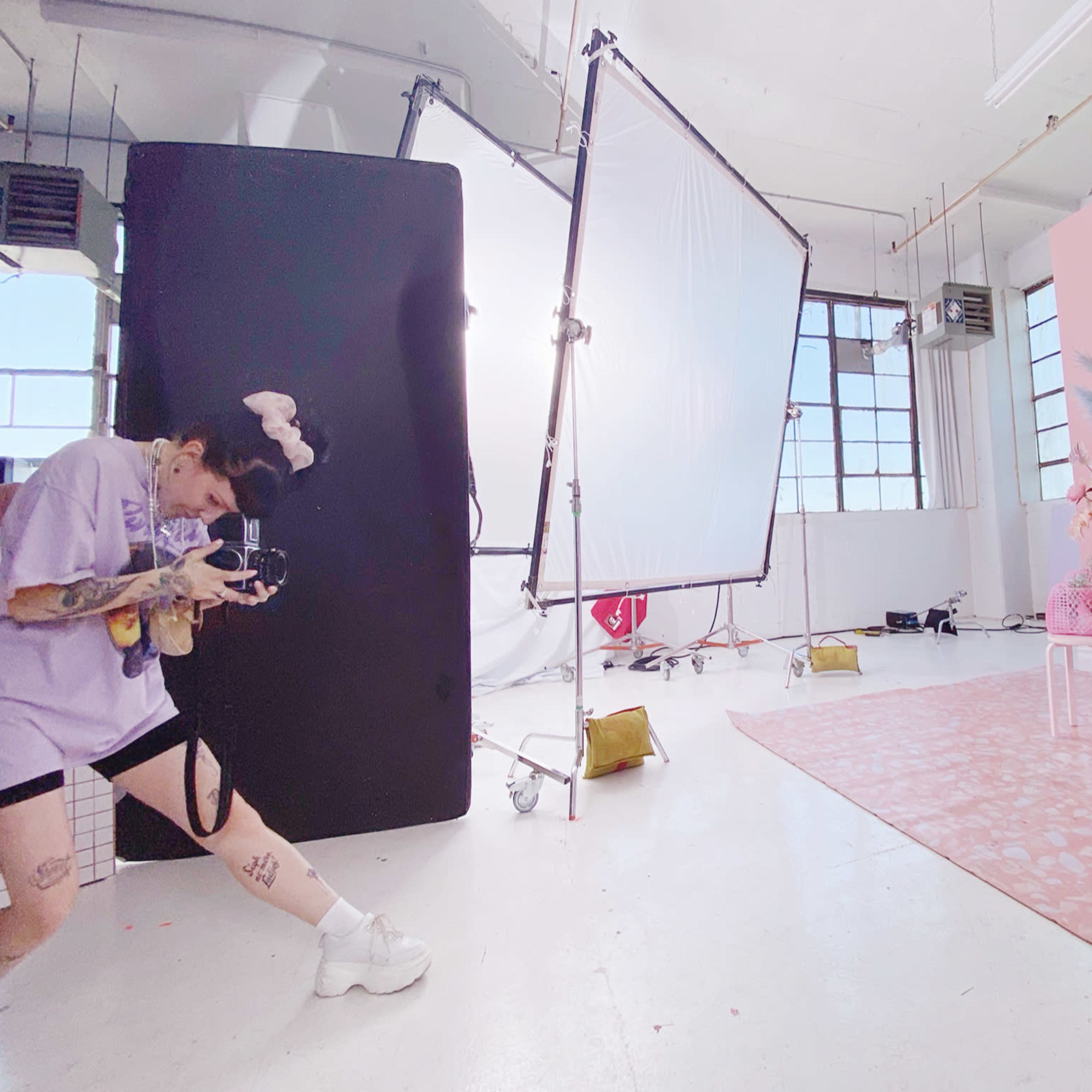 A photographer captures a model posing in a white dress against a backdrop of pink and floral decorations in a brightly lit studio.