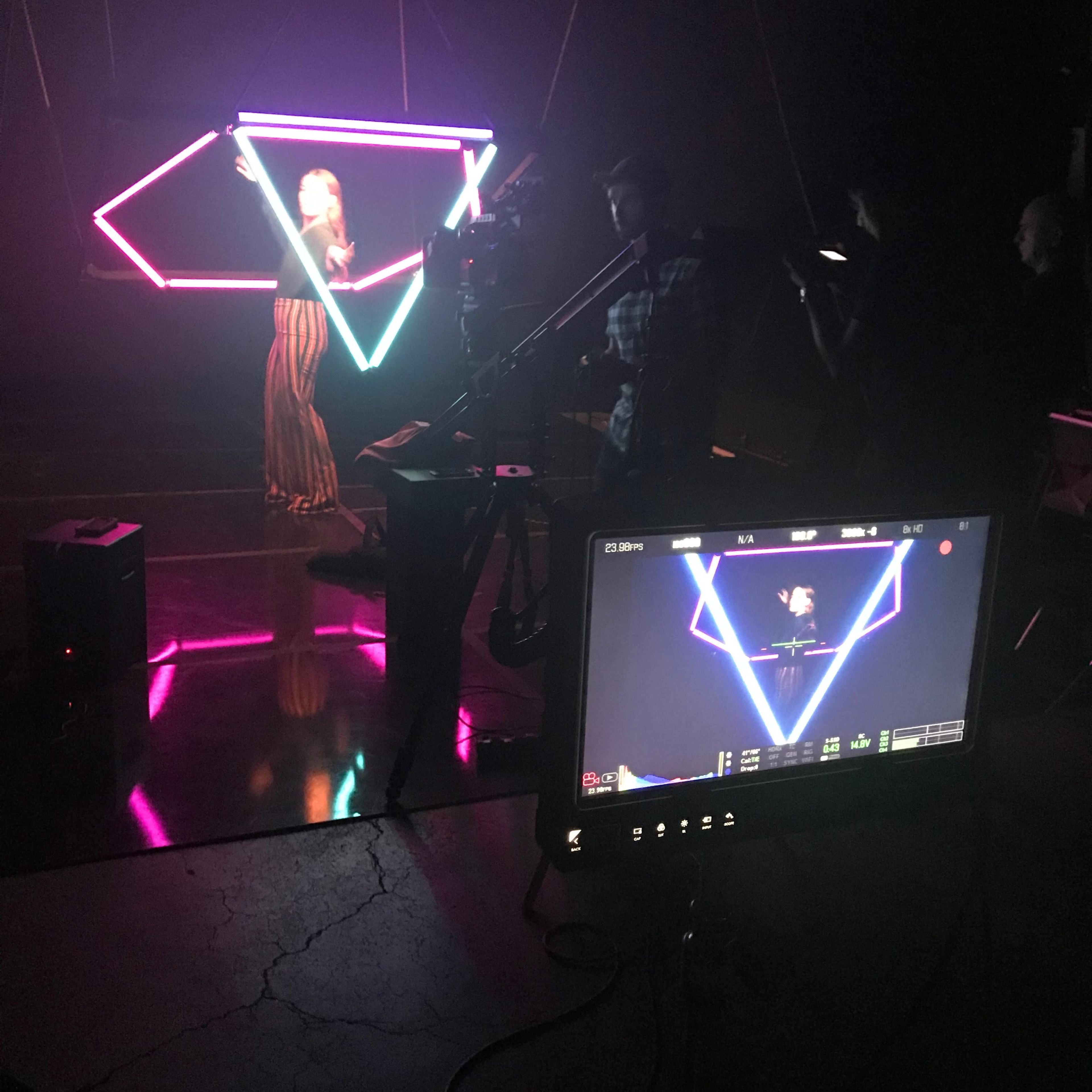 A person stands in front of a neon-lit geometric backdrop while a camera crew records from a monitor.