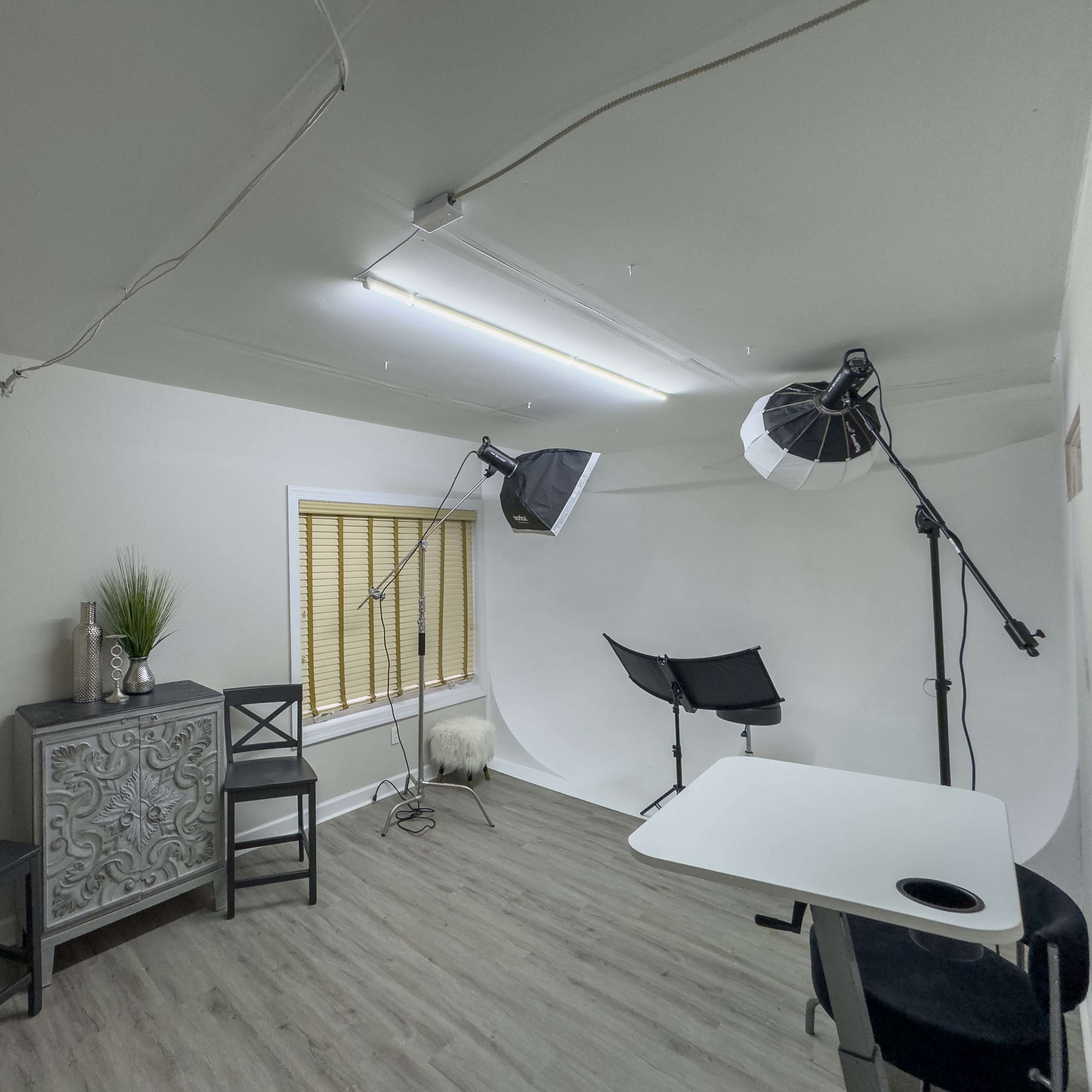 A small, well-lit studio space featuring a table, two chairs, a decorative cabinet, and photography equipment.