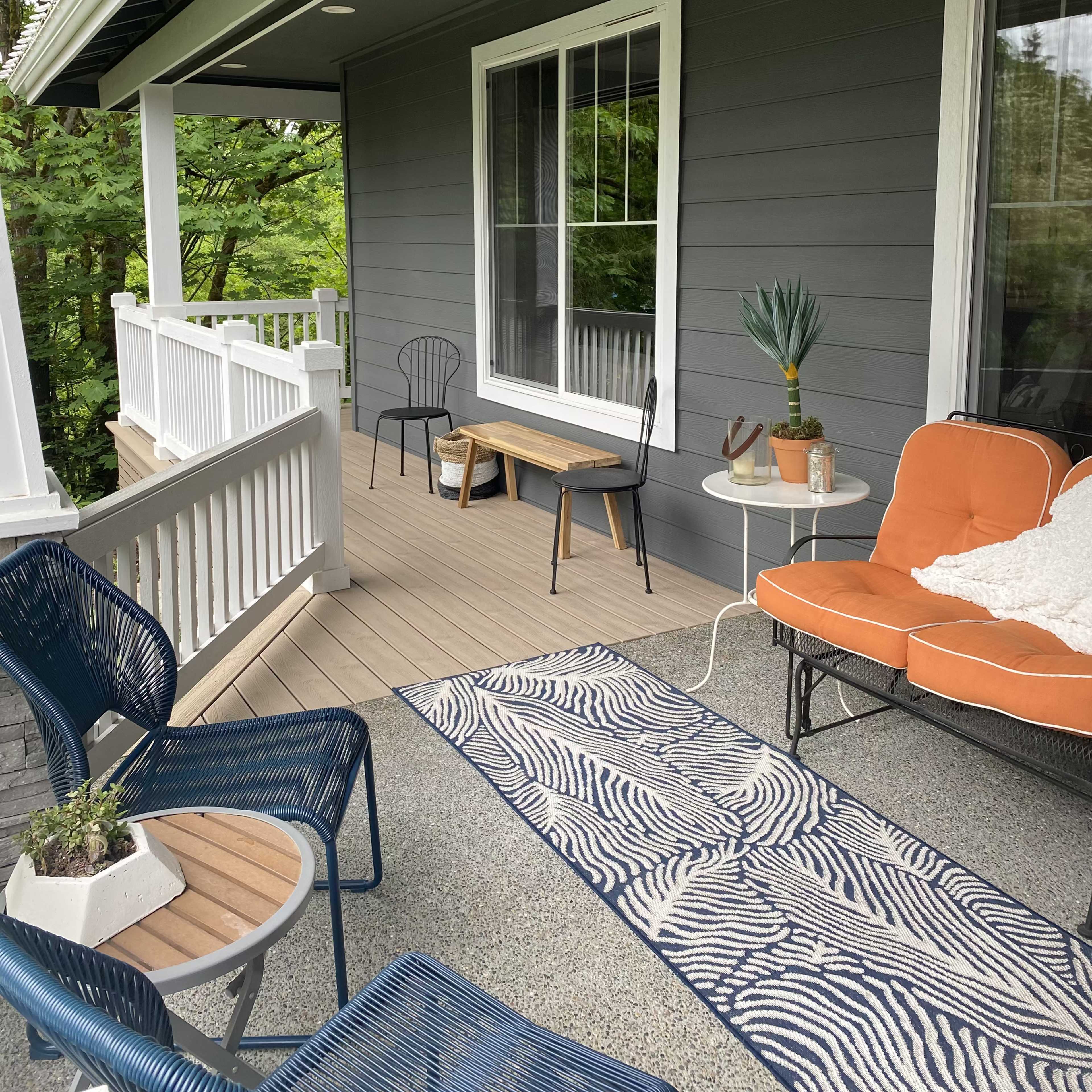 A covered porch with seating arrangements, including a bench, chairs, and a small table, surrounded by greenery.