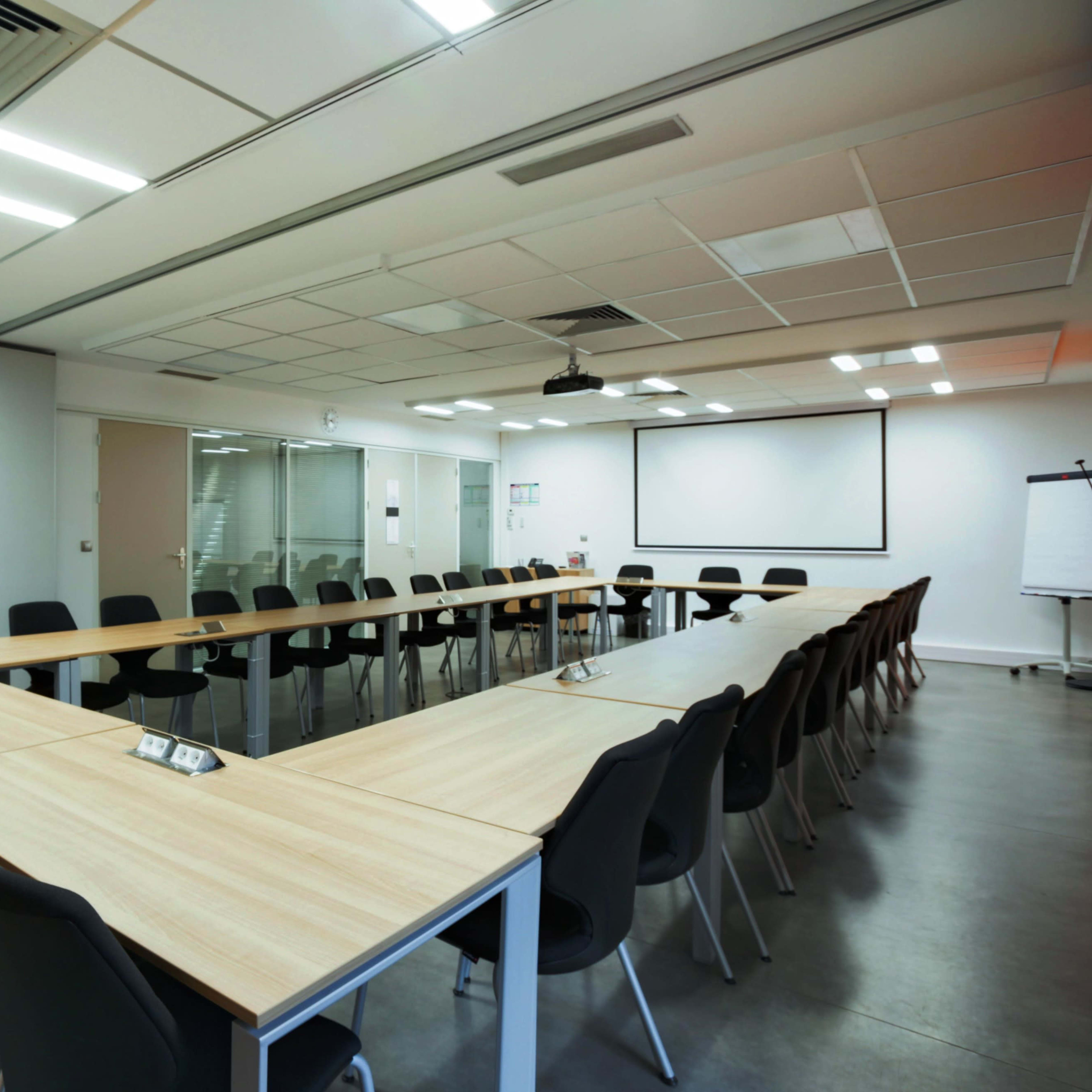 A large conference room features a U-shaped table surrounded by black chairs, with a projector screen at one end and glass walls.