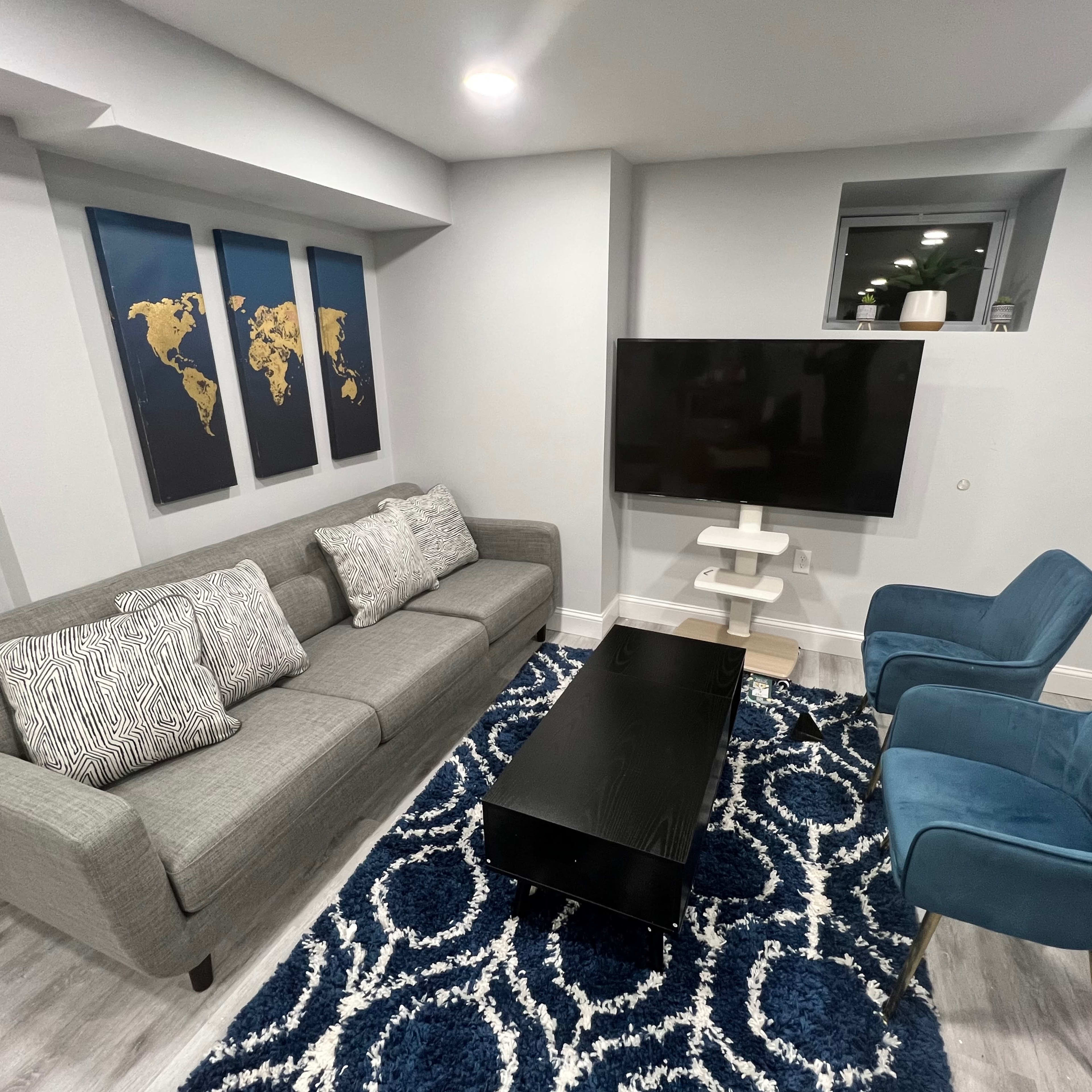 A modern living room featuring a gray sofa with decorative pillows, a black coffee table, two blue chairs, a TV mounted on the wall, and a world map art piece.