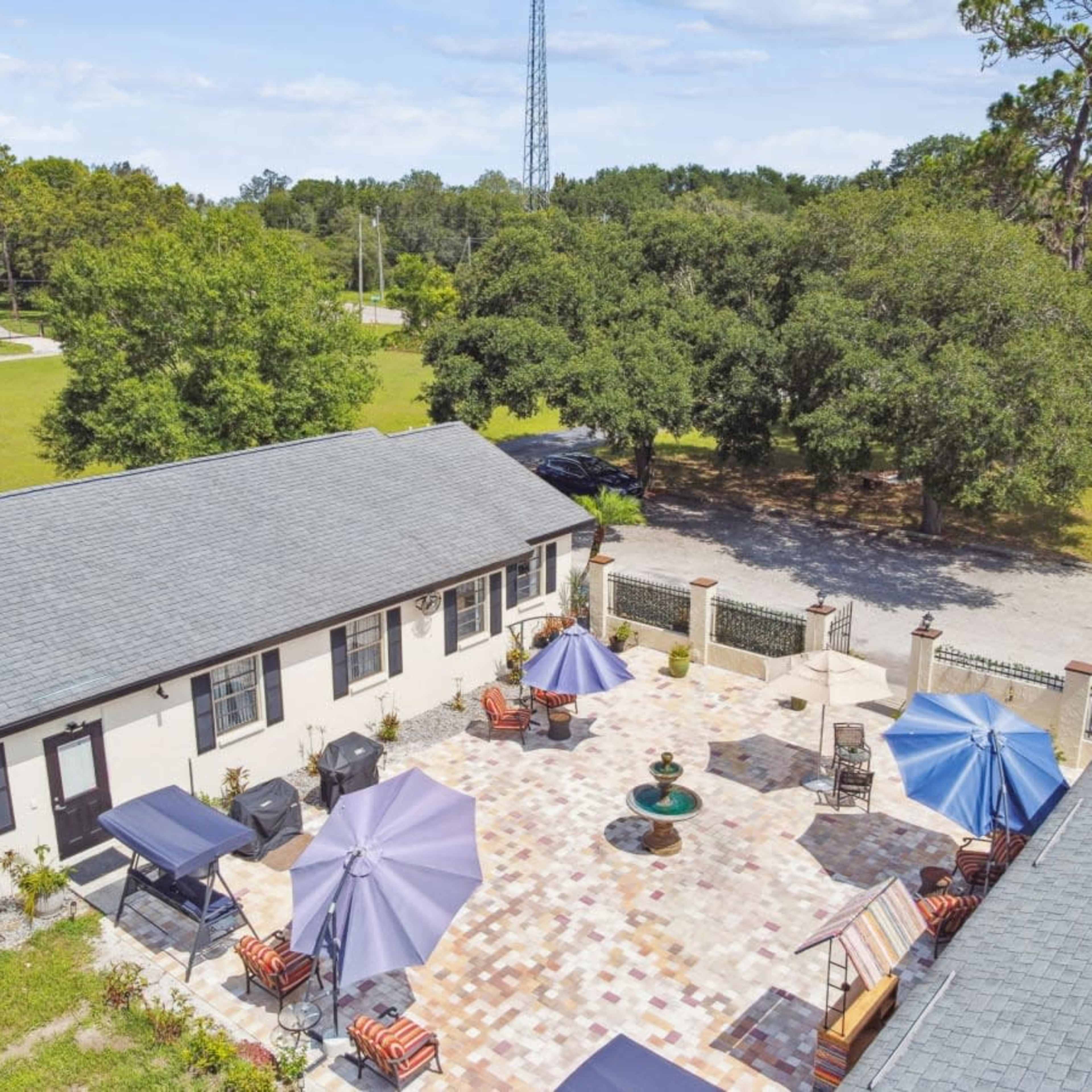 A spacious outdoor patio area with seating, umbrellas, and a fountain, surrounded by green trees and a clear sky.