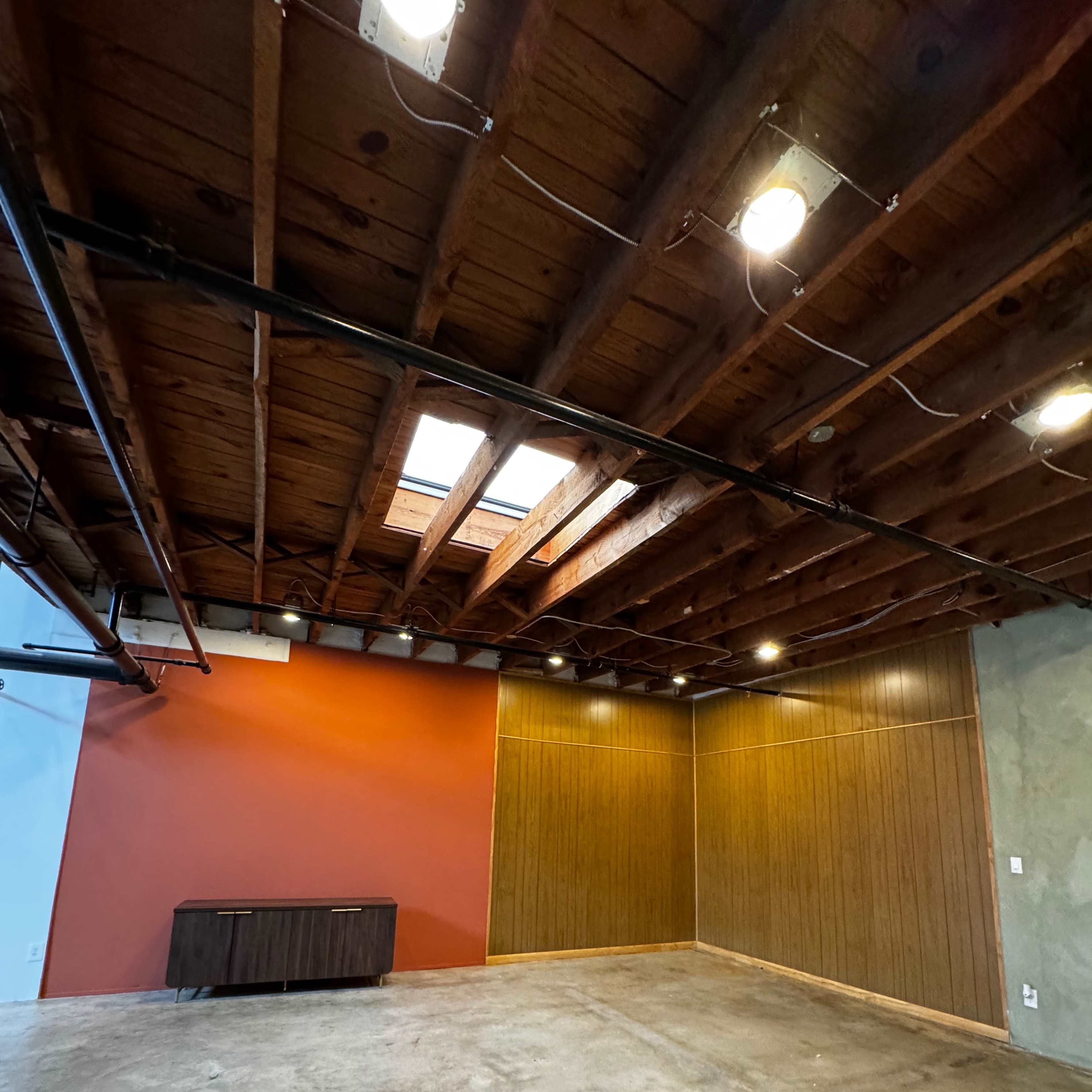 An empty room with wooden beams on the ceiling, a concrete floor, and one wall painted orange while another features wood paneling.
