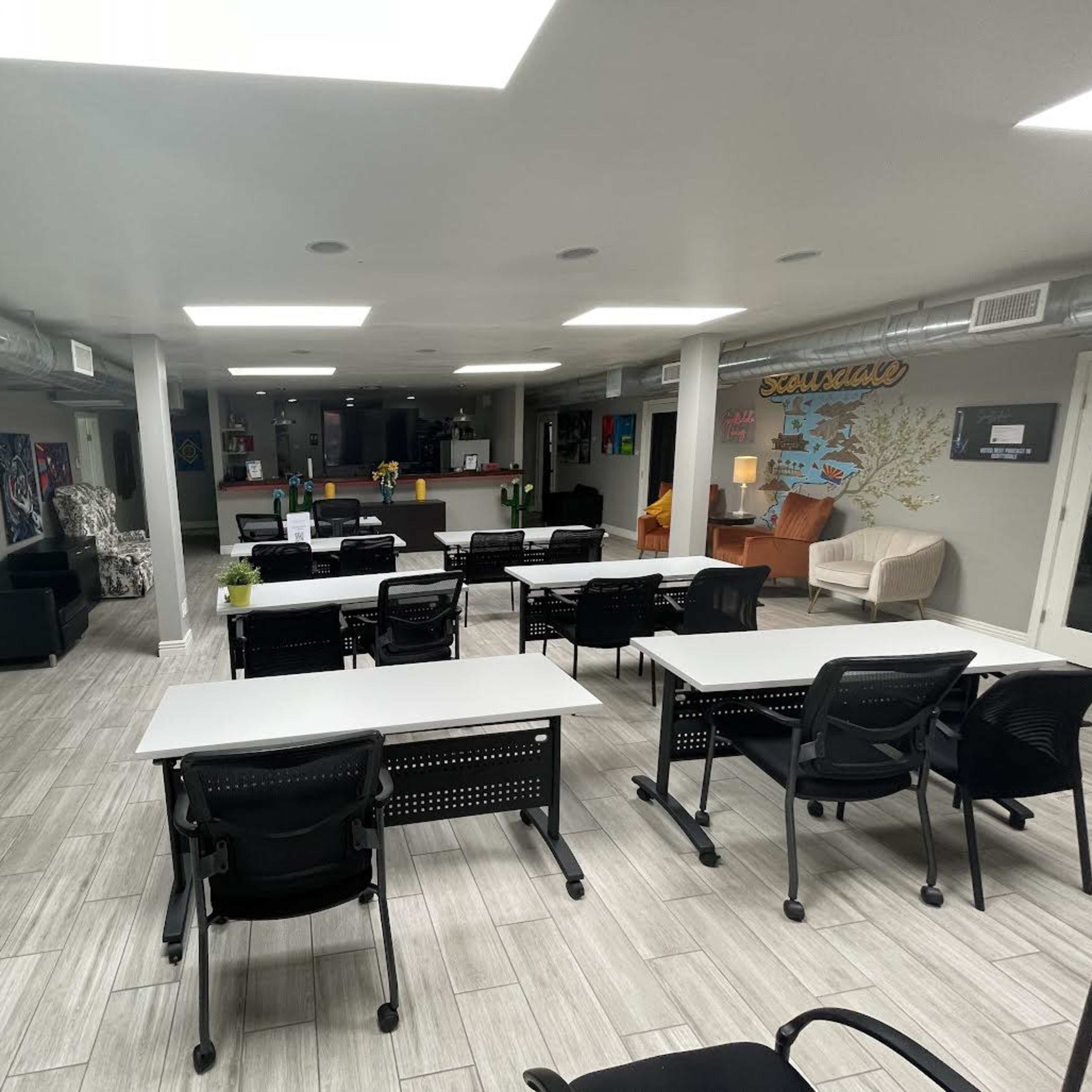 A spacious, modern office environment with multiple desks arranged for work, black chairs, and a lounge area featuring comfortable seating.