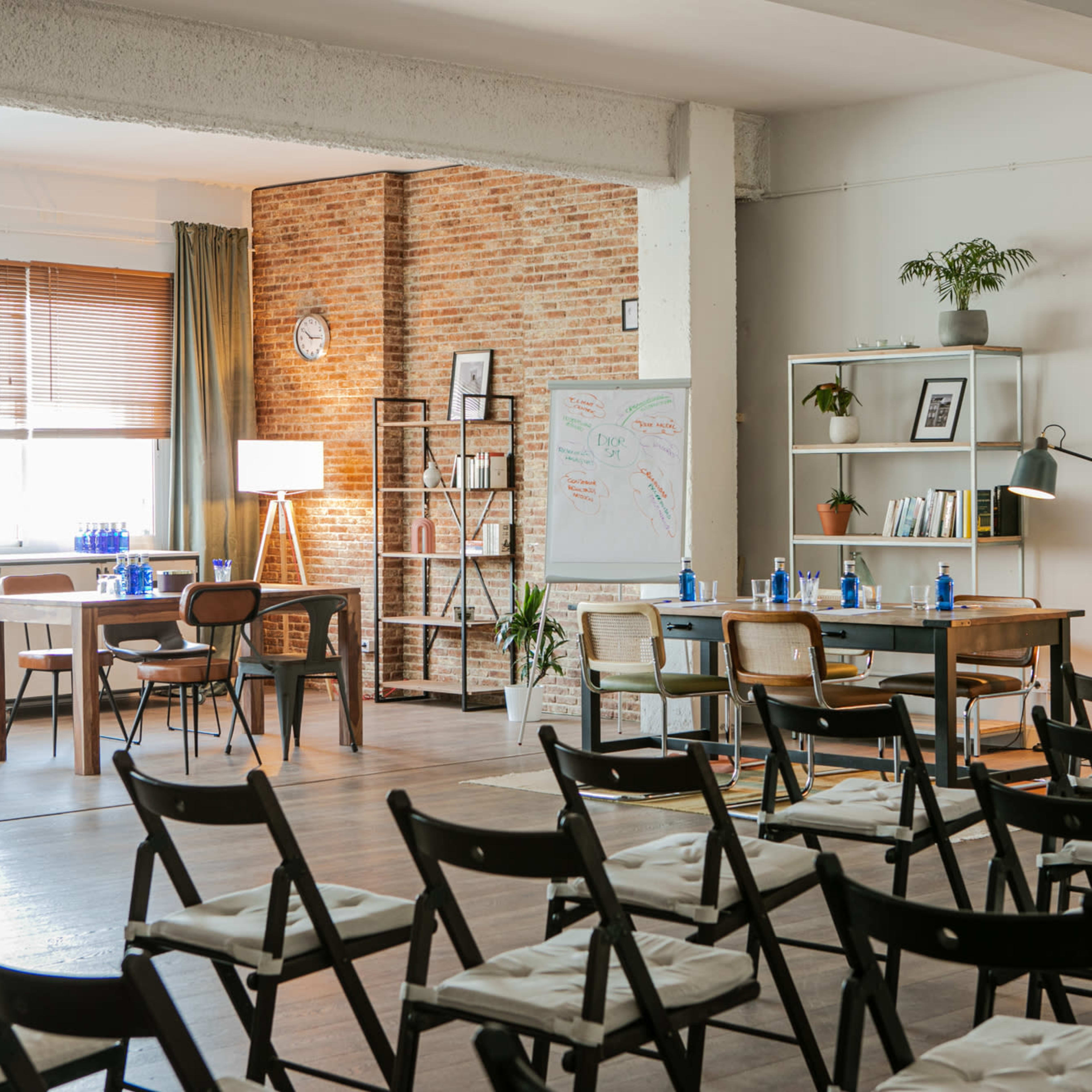 A modern meeting room with tables, chairs, a whiteboard, and plants, illuminated by natural light.