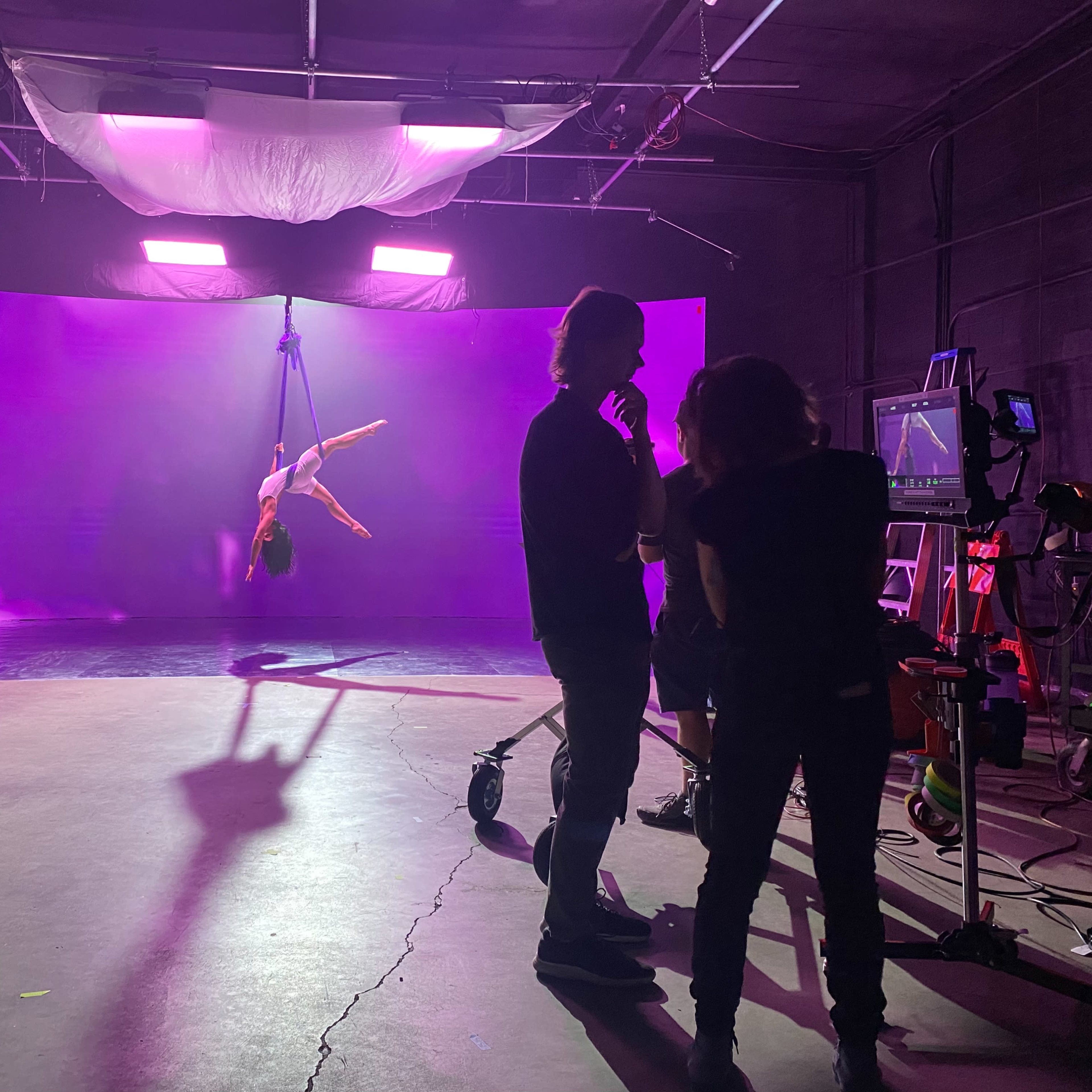 A performer is suspended from the ceiling on a harness against a luminous purple backdrop in a studio setting.