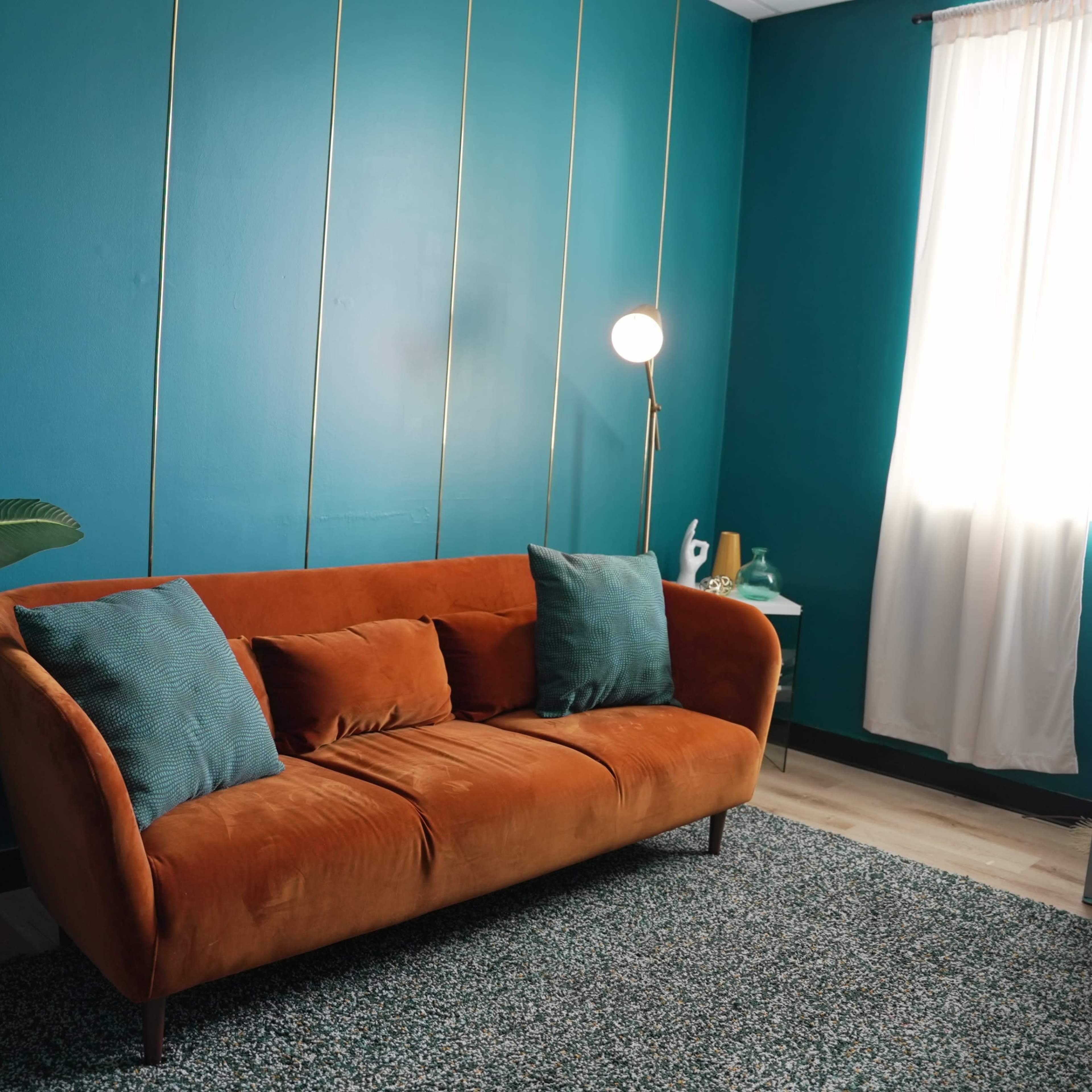 A modern living room featuring an orange velvet sofa, a large white planter with leaves, a floor lamp, and a desk beside a window with sheer curtains.