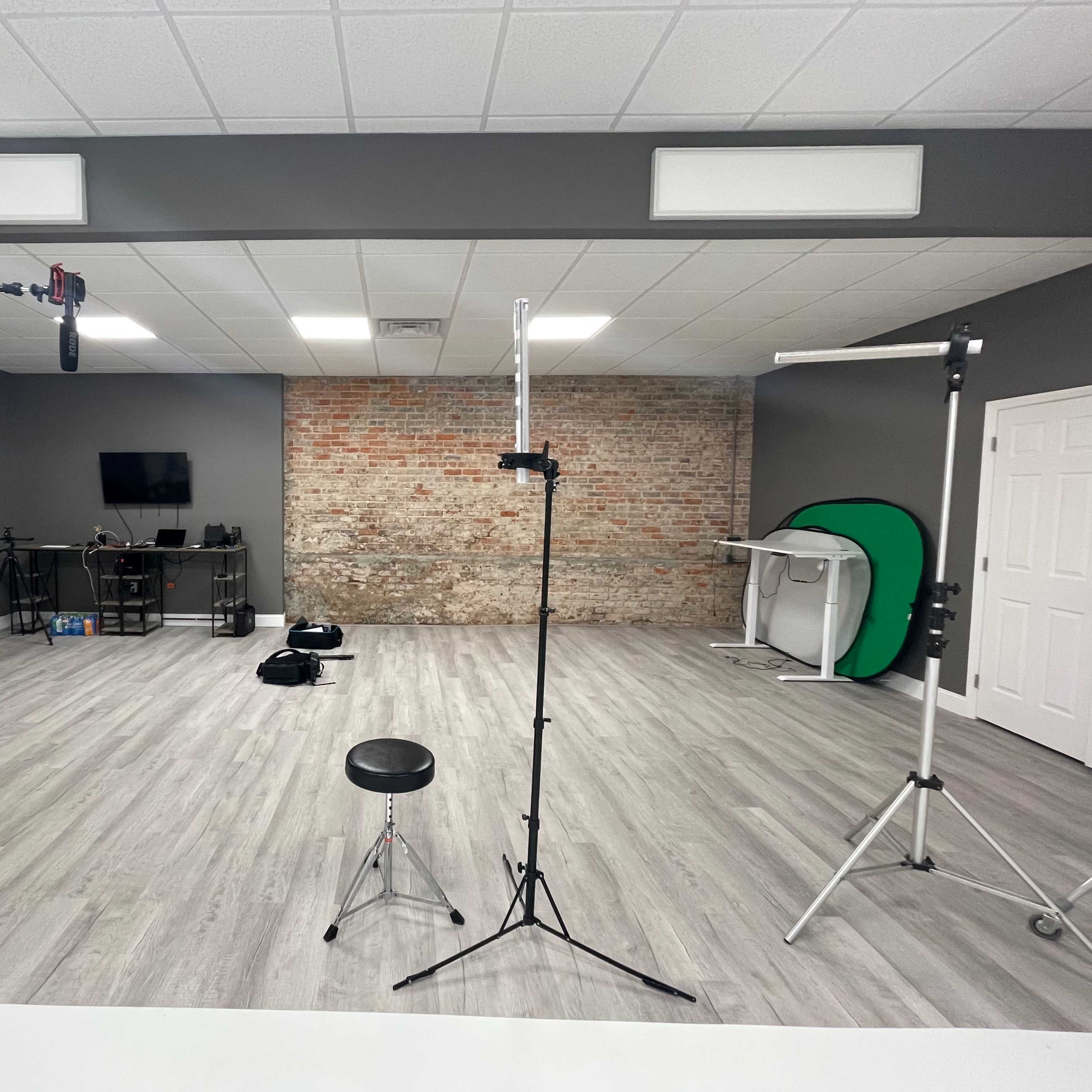 A spacious studio with wooden flooring, featuring several lighting stands, a backdrop area with a green screen, and equipment storage along a brick wall.