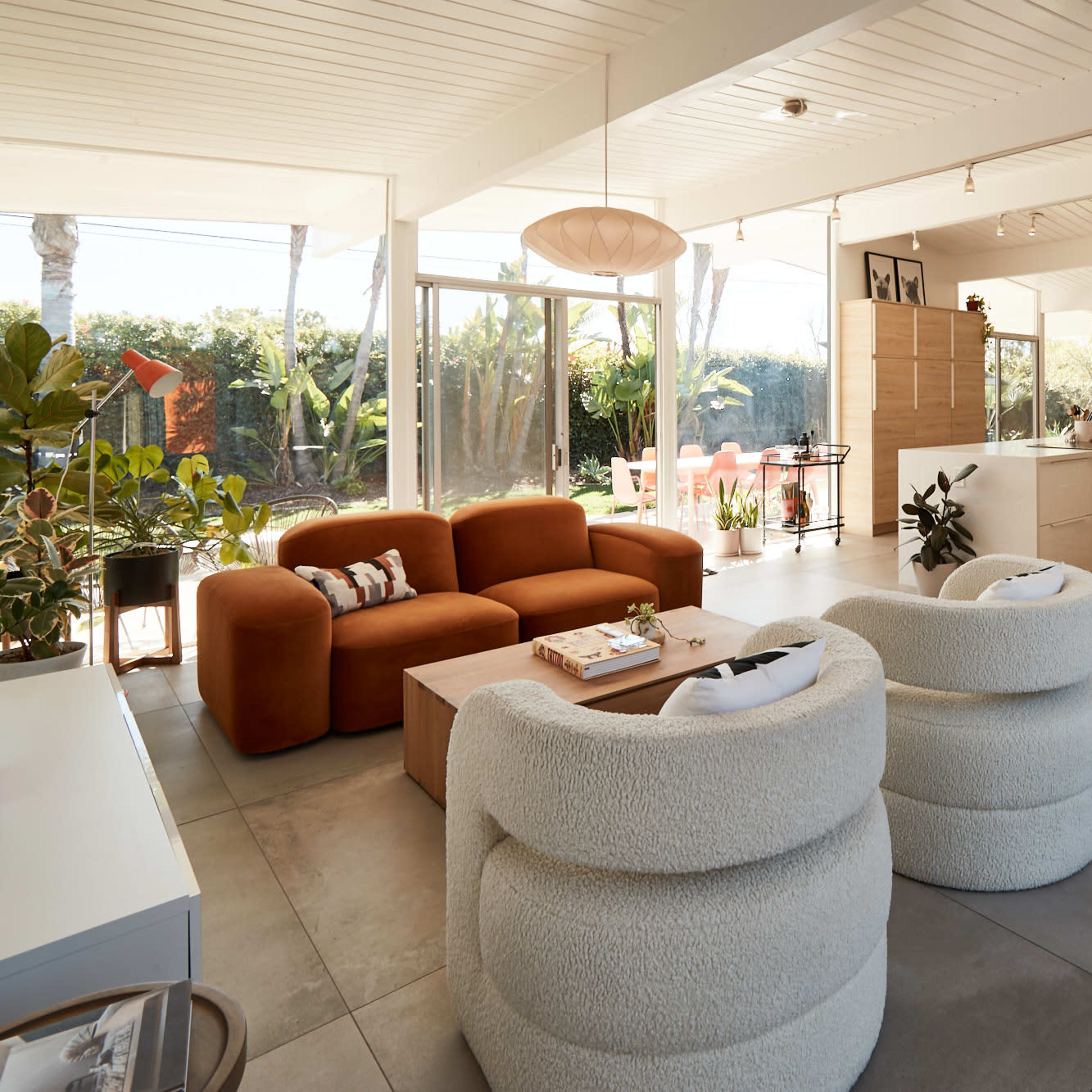 A modern living space with a brown sofa, two round white chairs, and a kitchen area visible in the background.