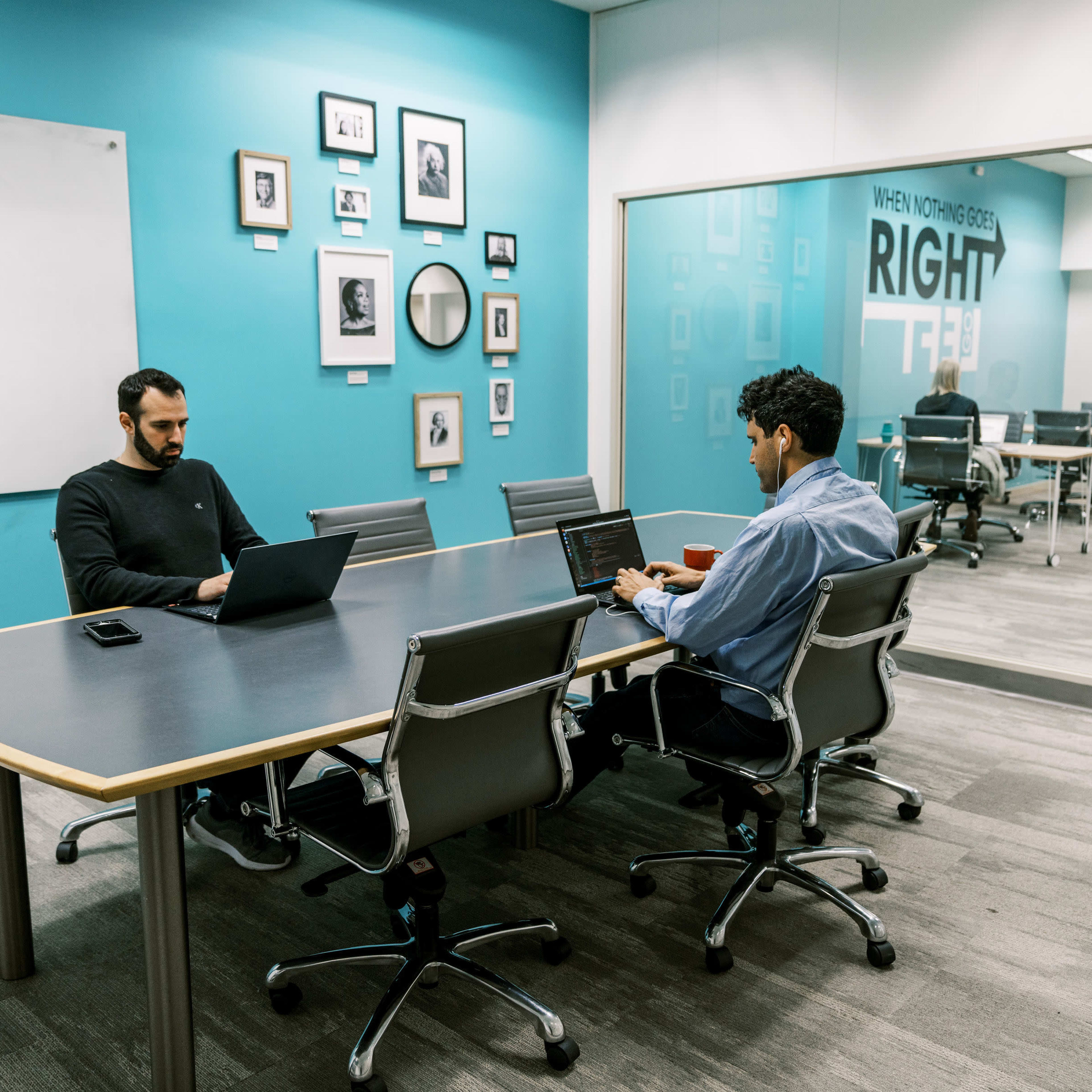 Two men are seated at a conference table, each working on laptops in a brightly colored meeting room.