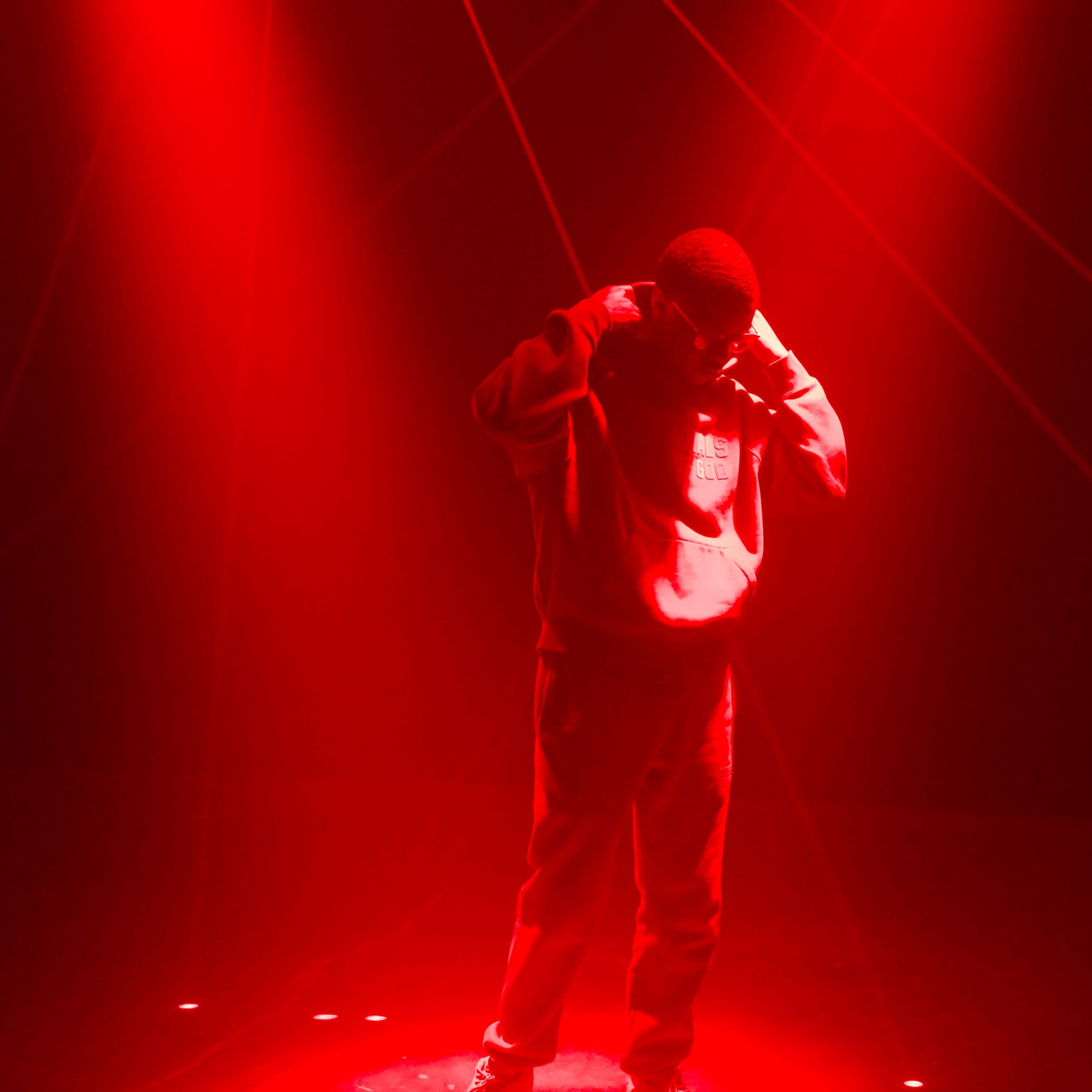 A person in a hoodie stands on a spotlighted stage under red lighting with laser lines crossing the background.