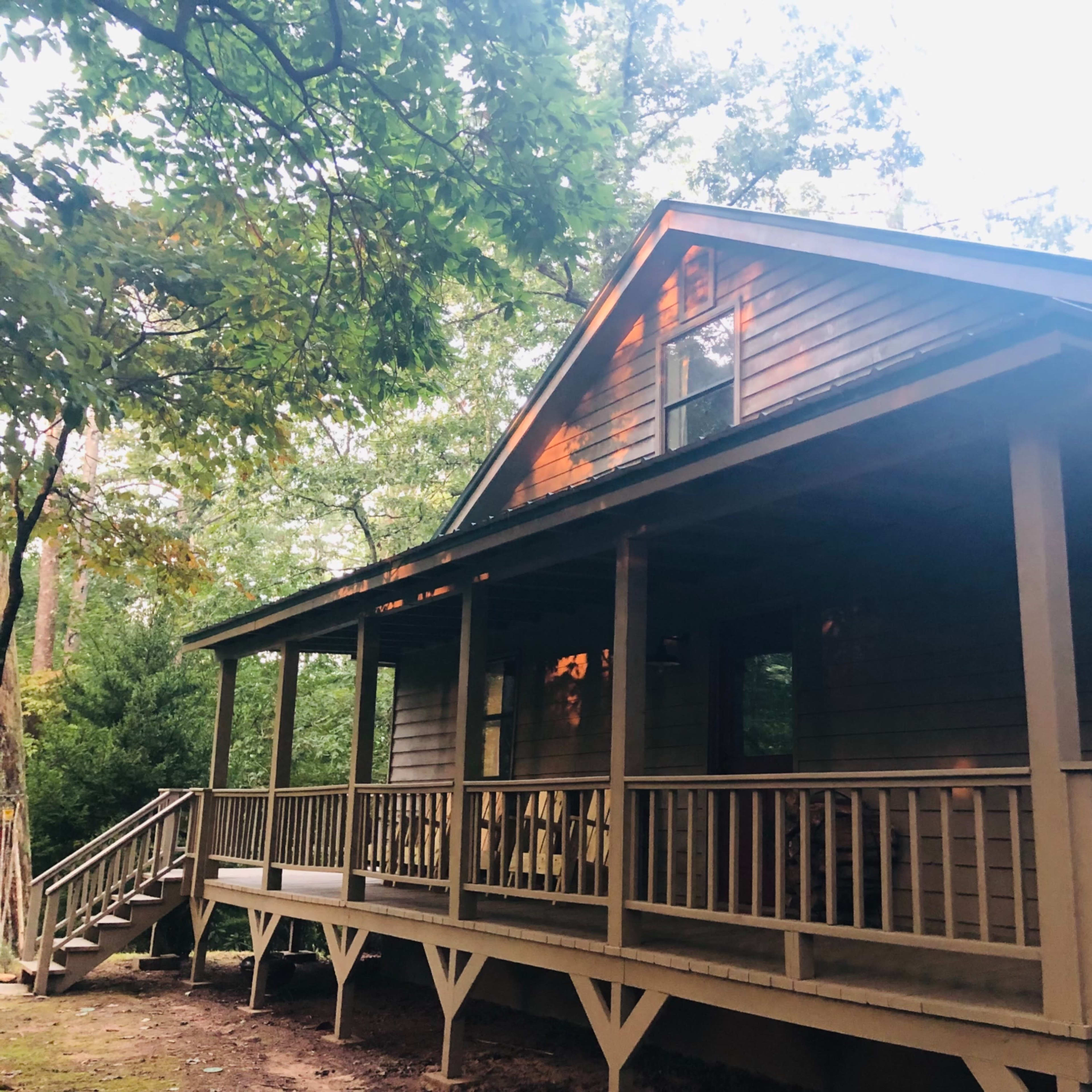 A wooden house with a front porch sits among trees in a wooded area.