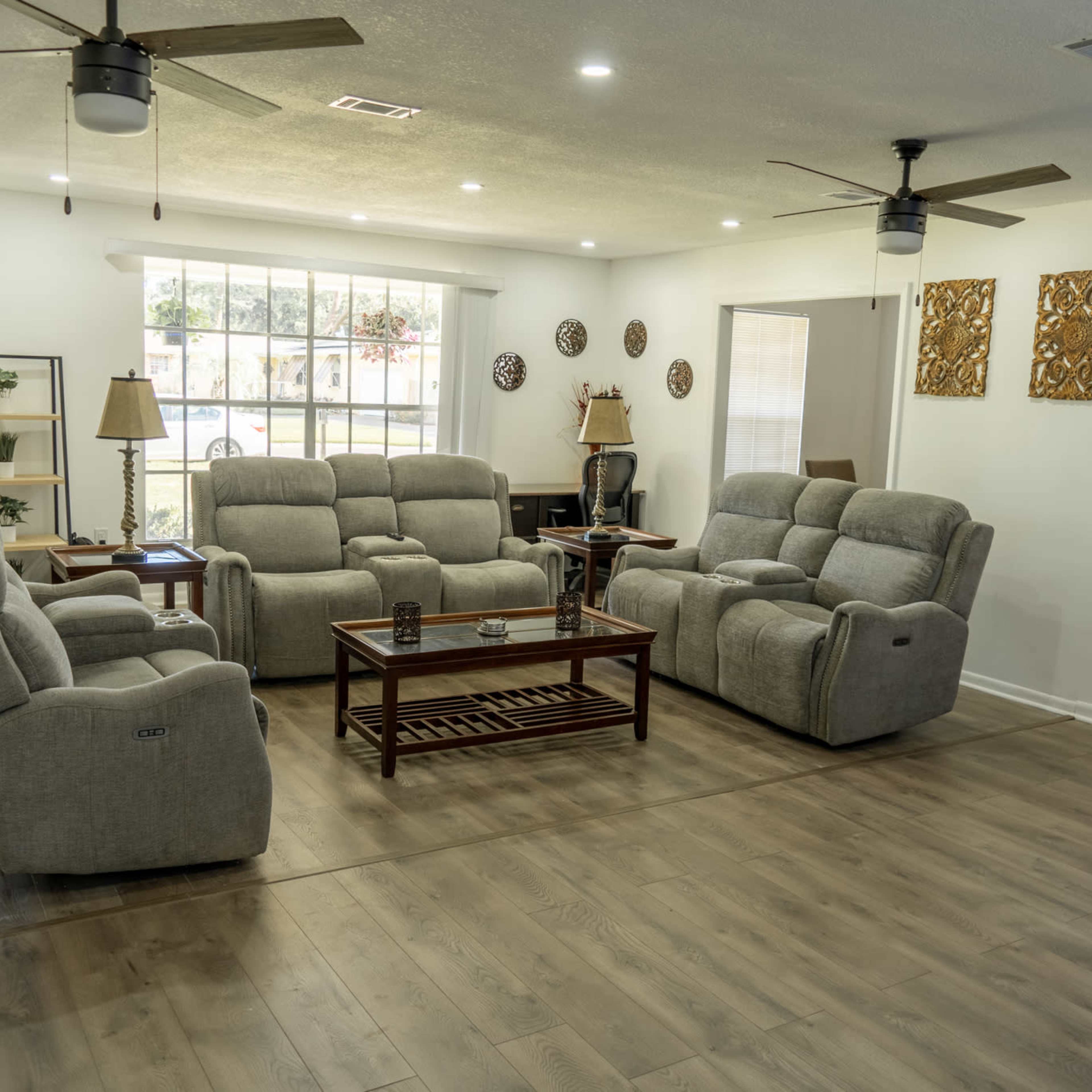 The room features a spacious layout with gray reclined sofas arranged around a wooden coffee table, set in front of a large window and a light-colored front door.