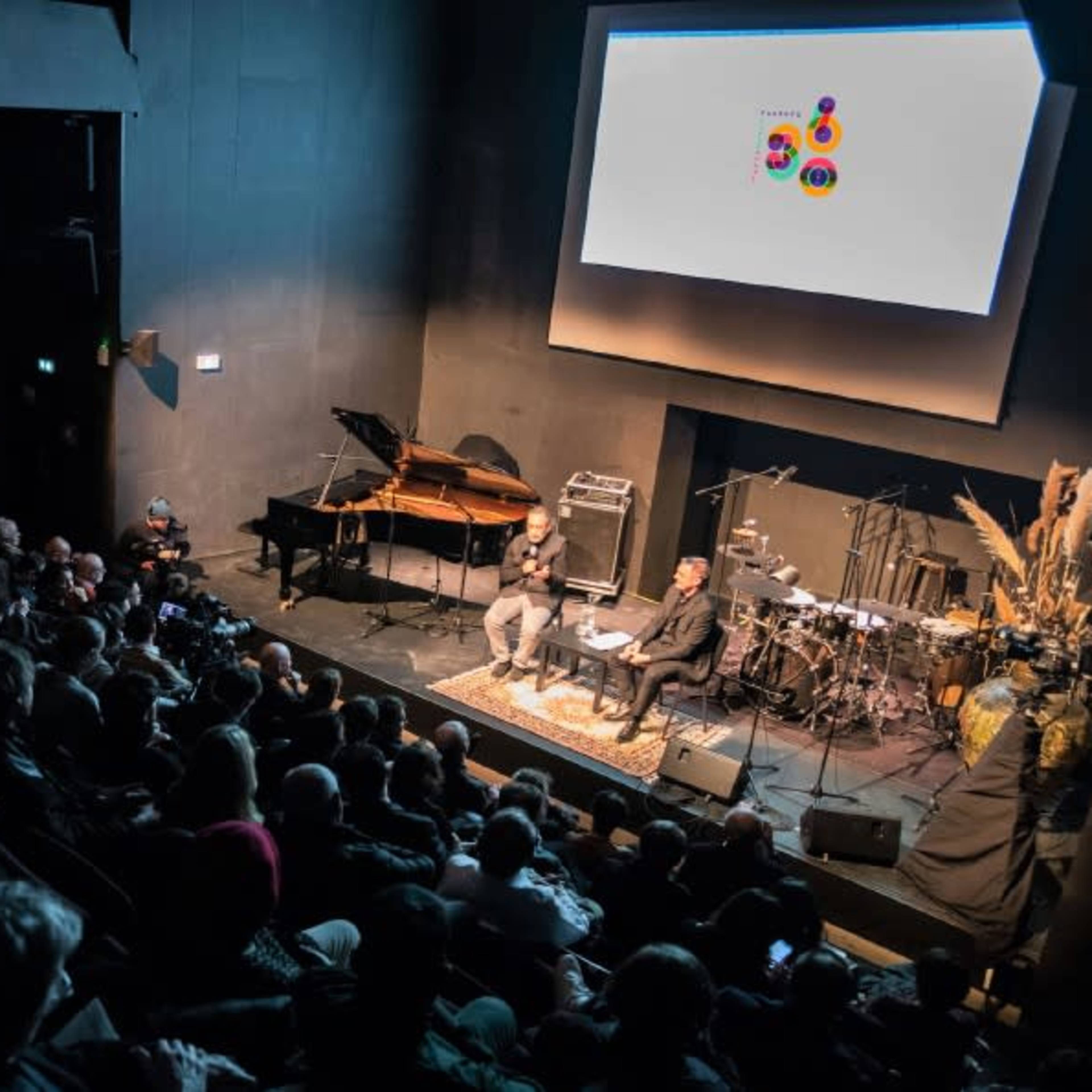 A large audience watches a discussion on stage with a piano and a band setup in the background.
