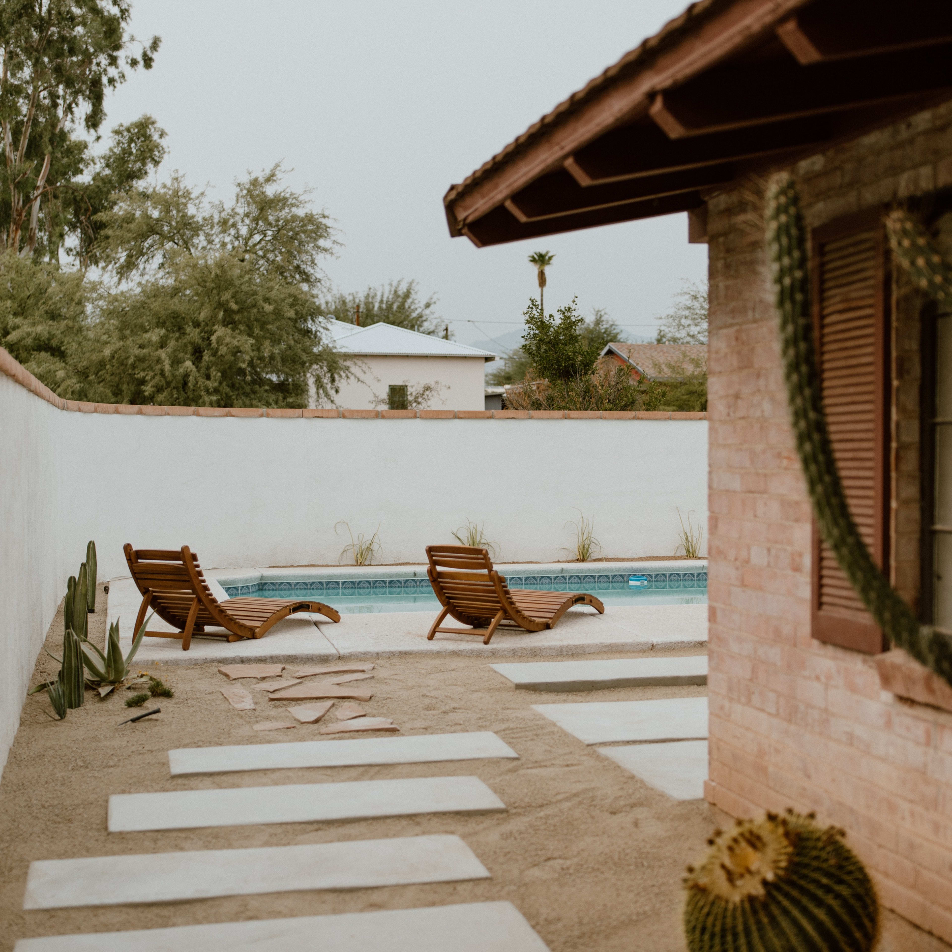 A view of a pool area featuring two lounge chairs, surrounded by a sandy landscape with cacti and stepping stones.