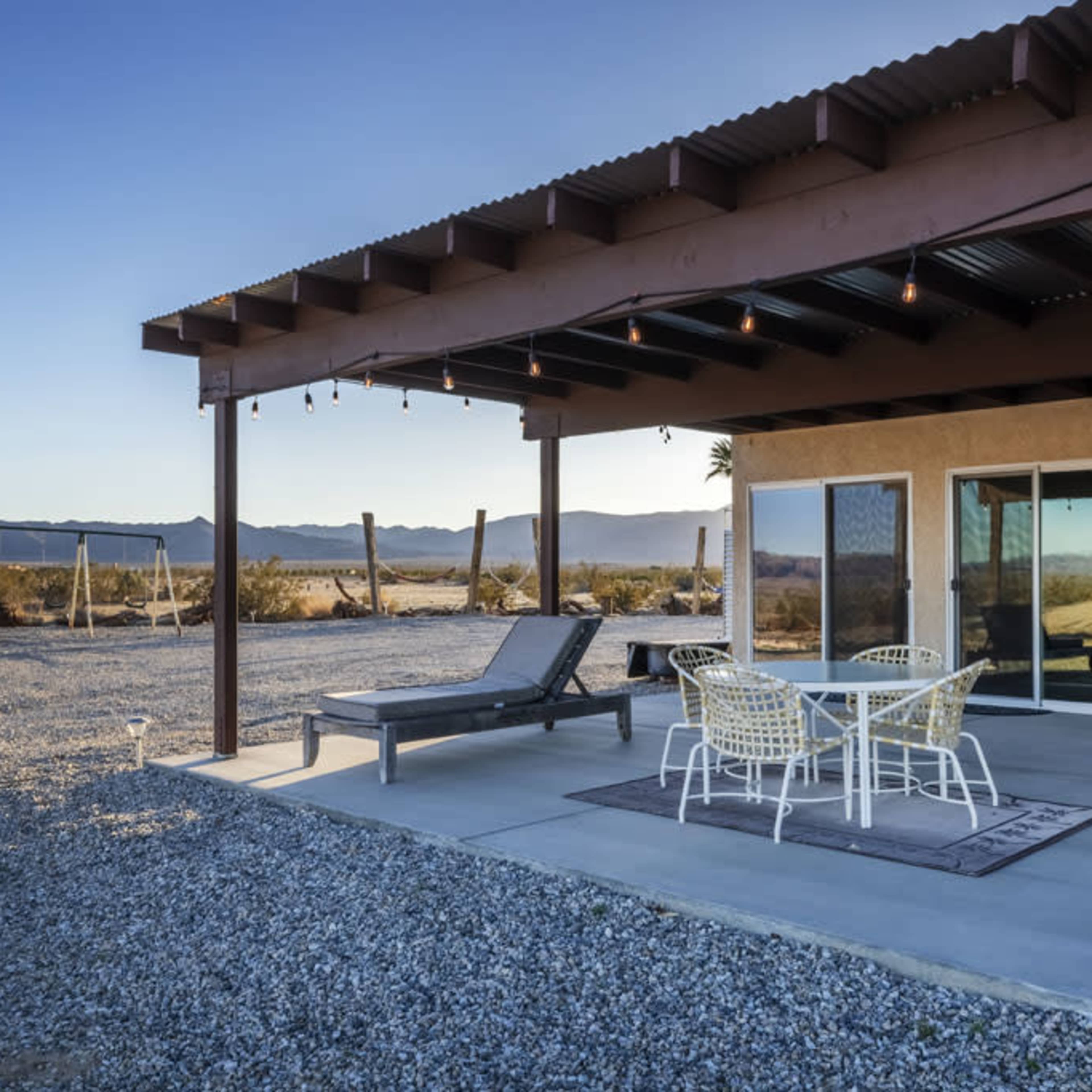 A patio area of a house with a table and chairs, surrounded by gravel and mountain scenery in the background.