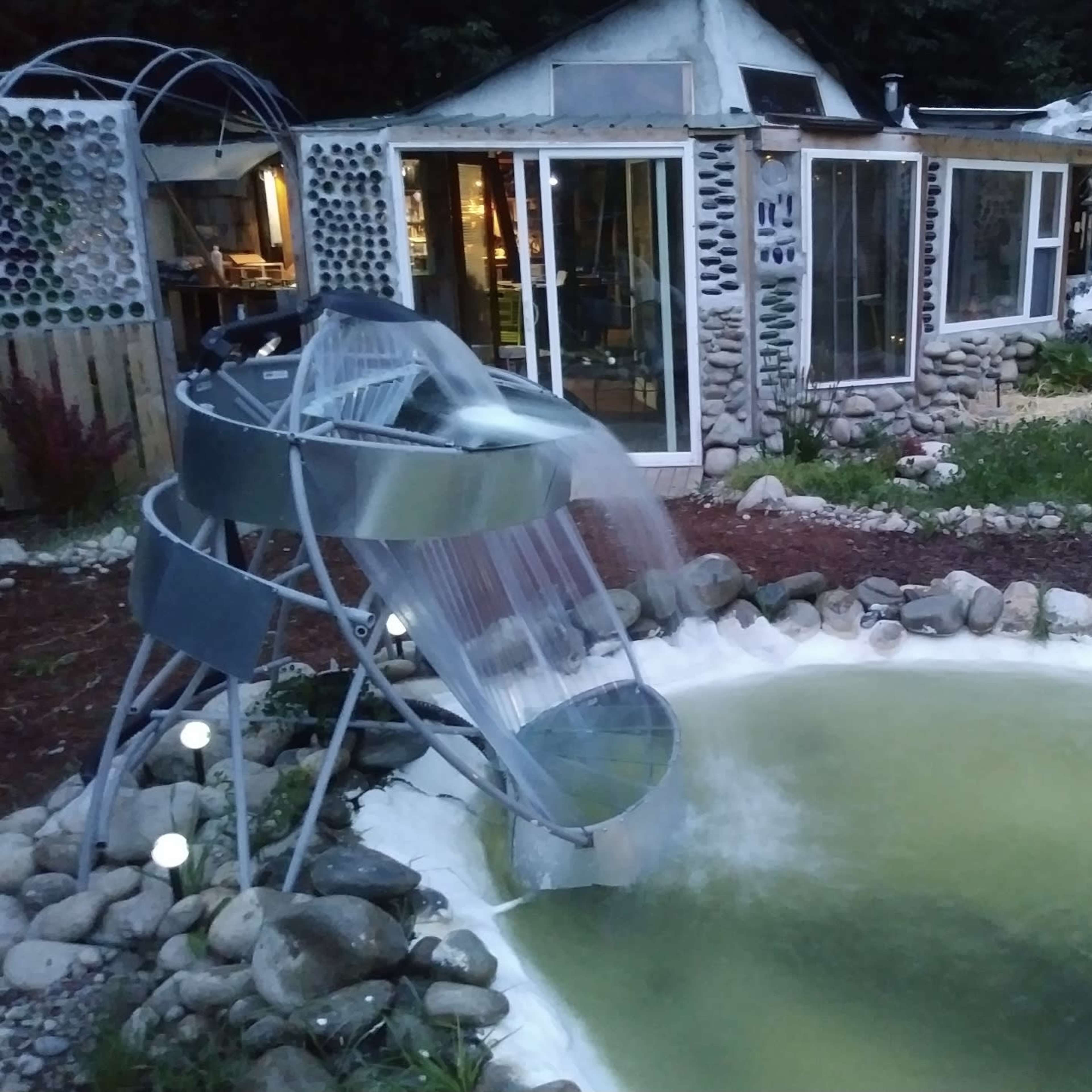 A modern water slide spills into a small, green pond surrounded by smooth stones and a rustic greenhouse in the background.