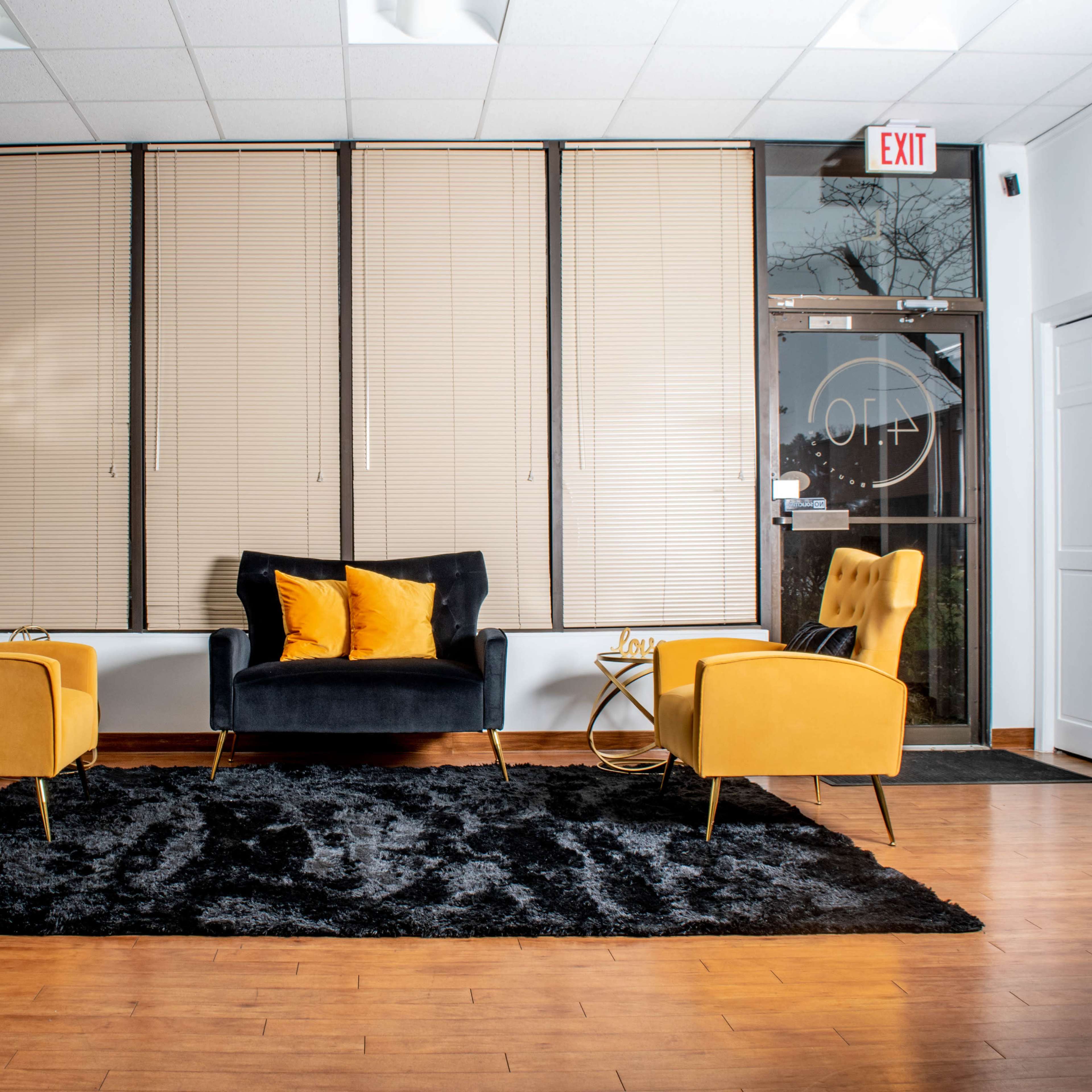 A modern waiting room featuring two yellow chairs, a black sofa, a coffee table, and a large black rug, with large windows covered by blinds.