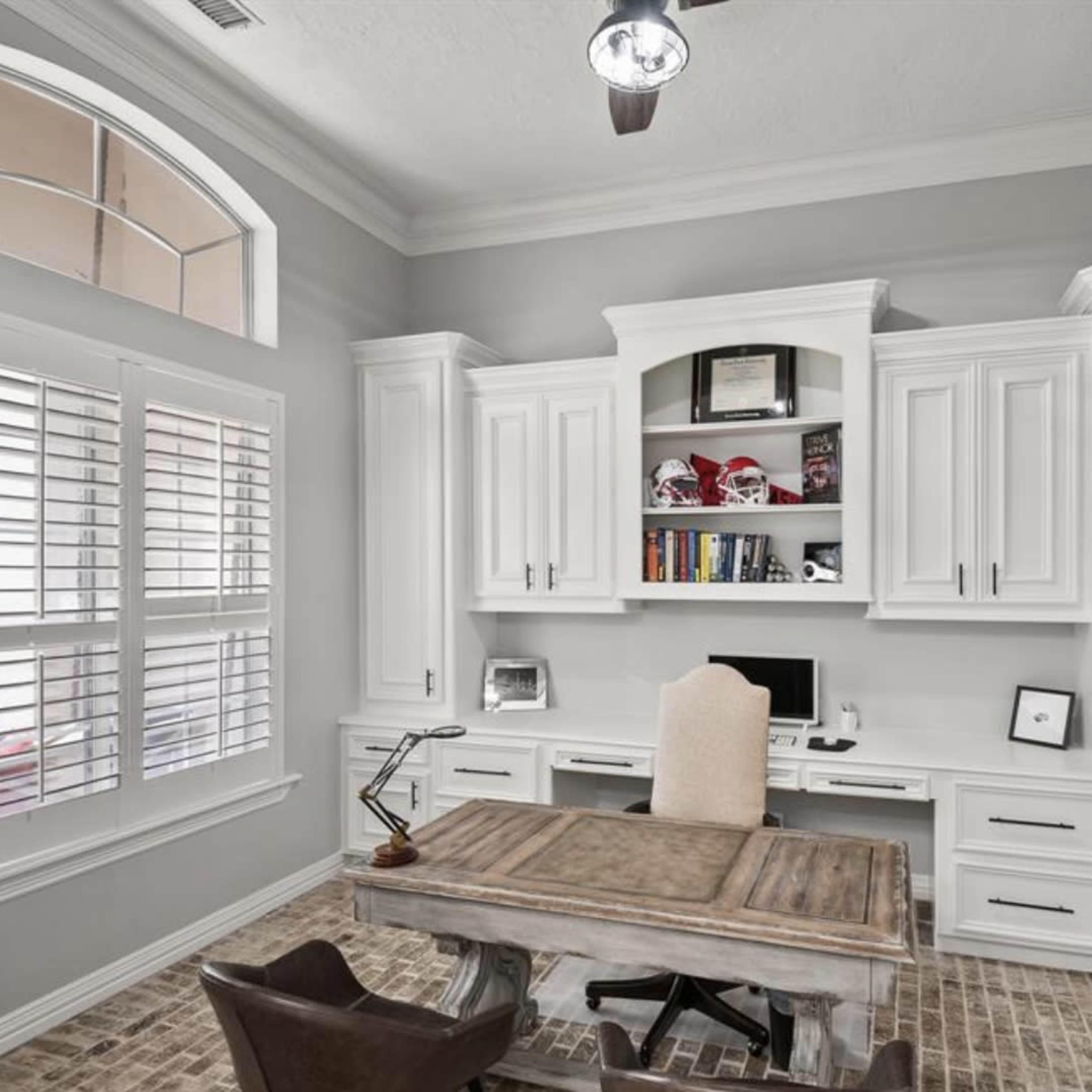 A home office with a wooden desk, white cabinetry, and large windows with plantation shutters.