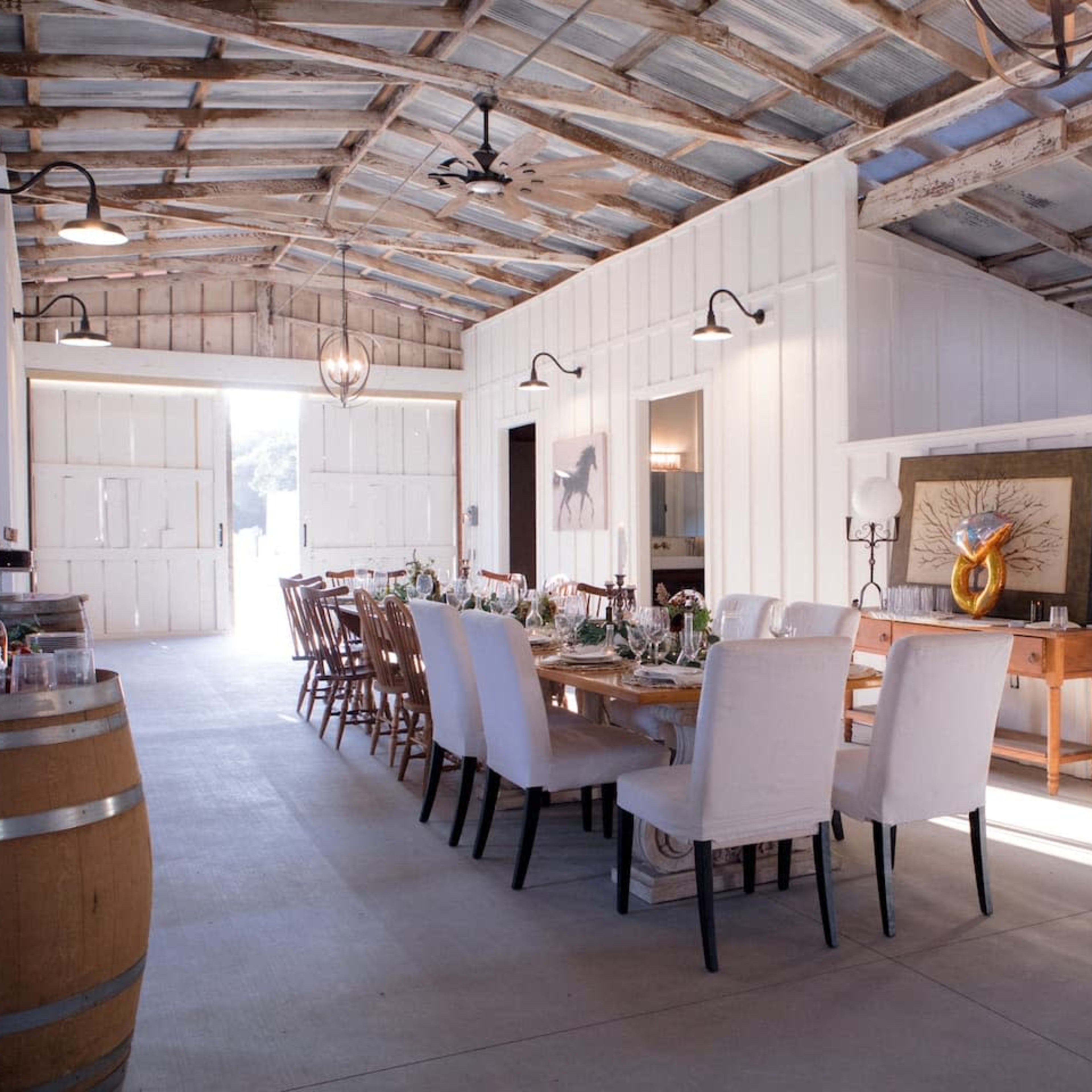 A spacious, rustic dining room with long wooden tables set for a meal, surrounded by white chairs, within a building featuring exposed beams and large double doors.