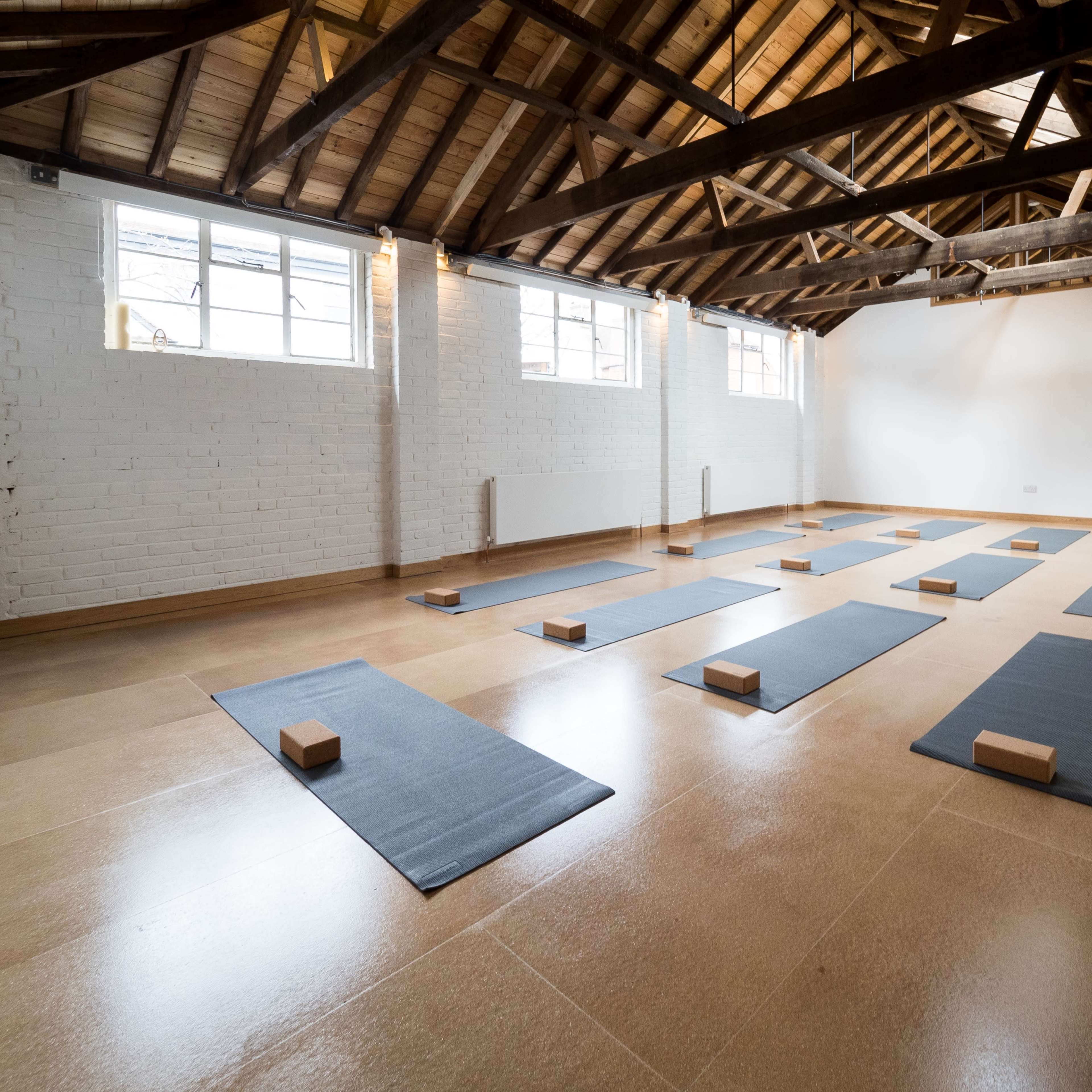 A spacious yoga studio features neatly arranged mats and wooden blocks on the floor beneath a high, wooden-beamed ceiling.