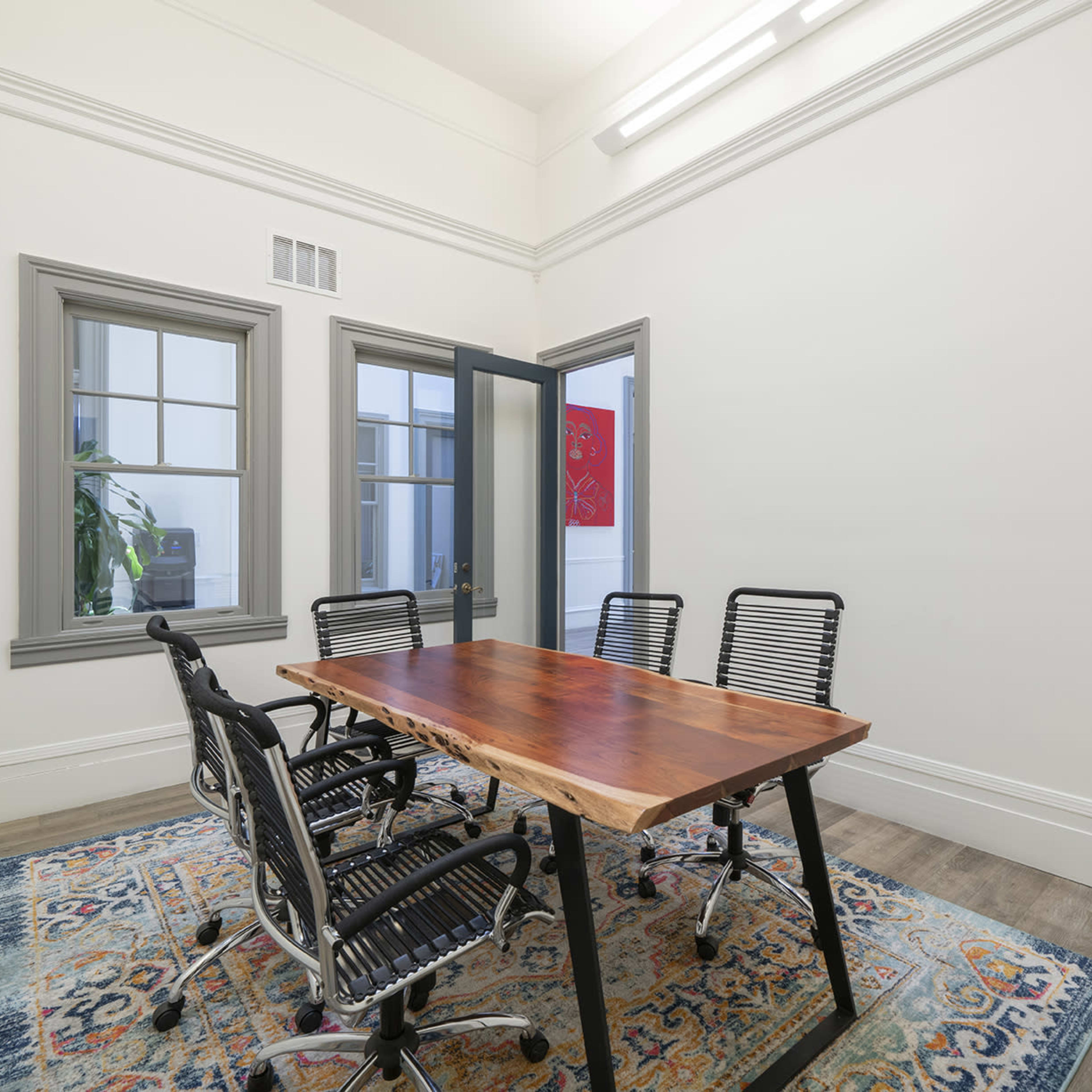 A conference room features a wooden table surrounded by five black office chairs, with large windows and a patterned rug on the floor.