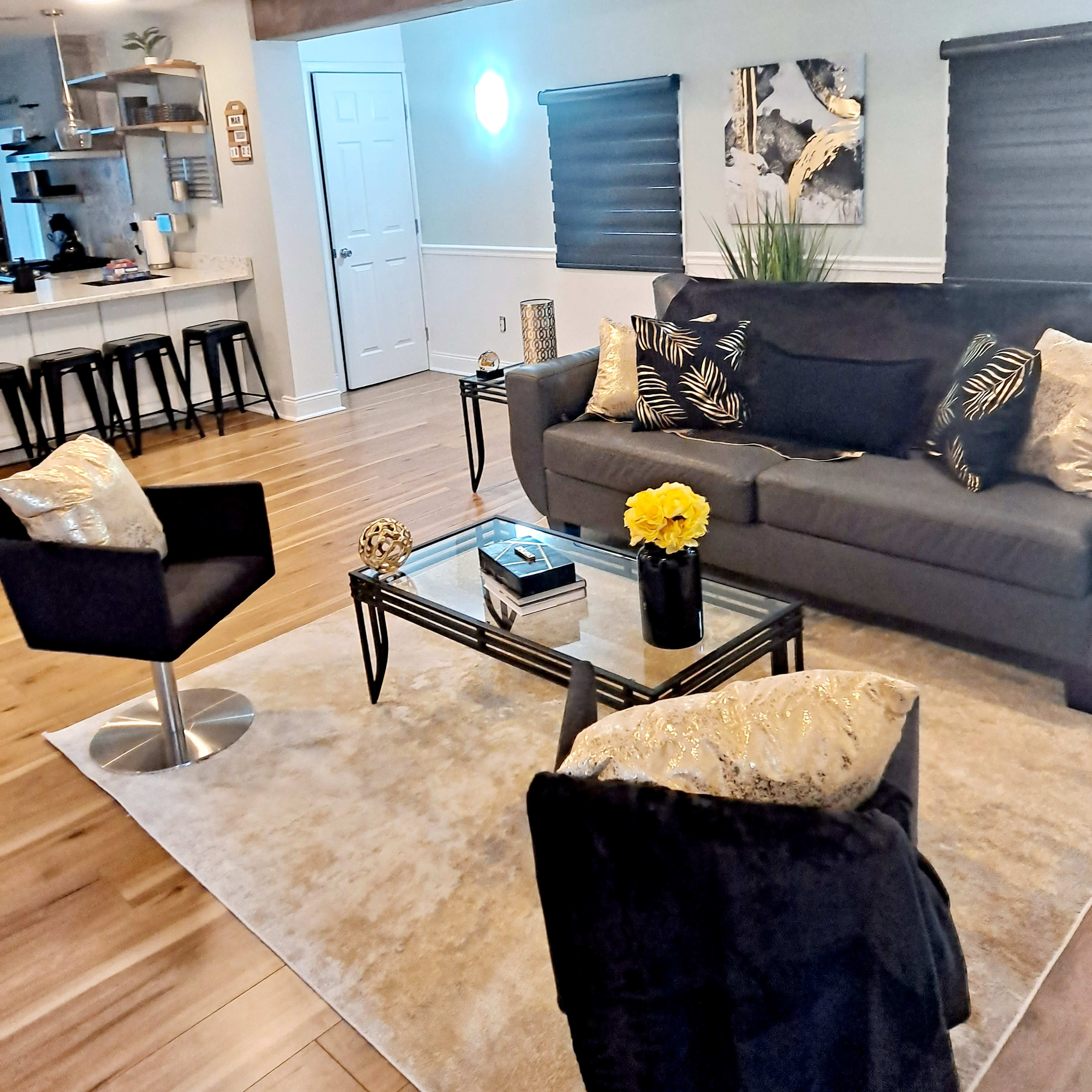 A modern living room features a gray sofa, black chairs, a glass coffee table, and a decorative rug, with open sightlines to a kitchen area.