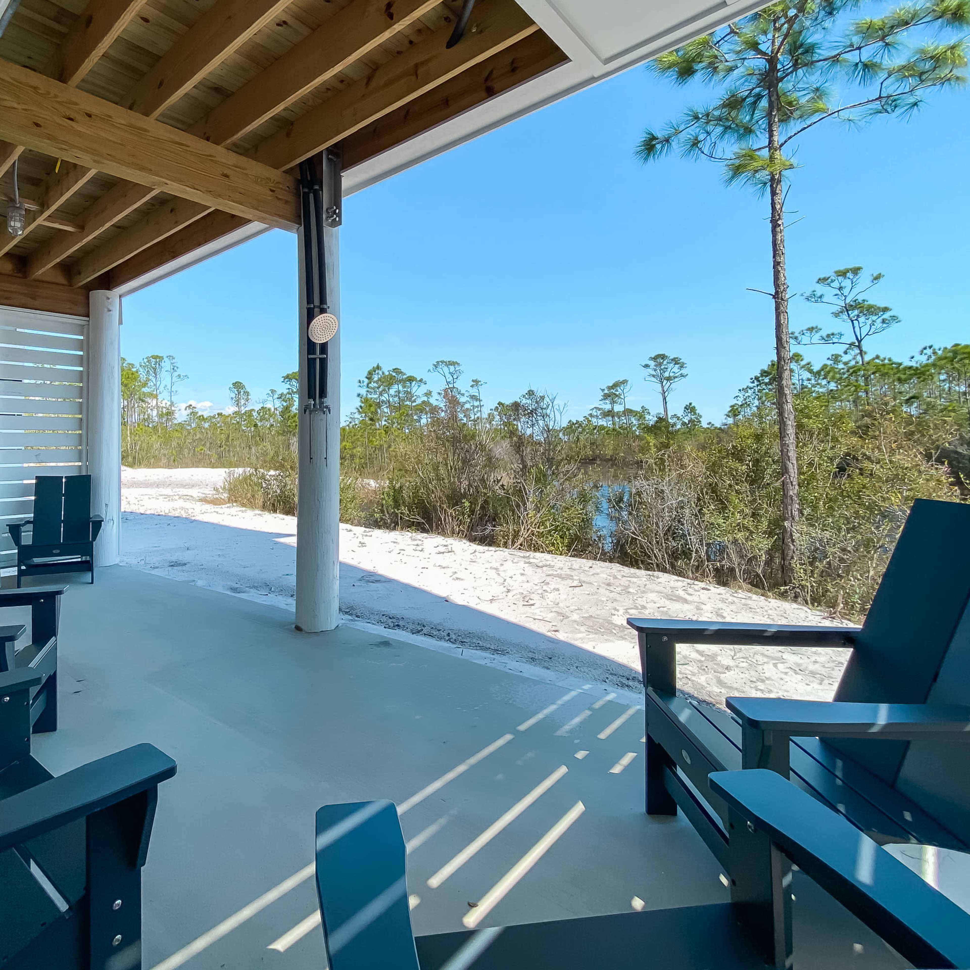 A covered patio with blue Adirondack chairs overlooks a sandy area and a body of water surrounded by vegetation.