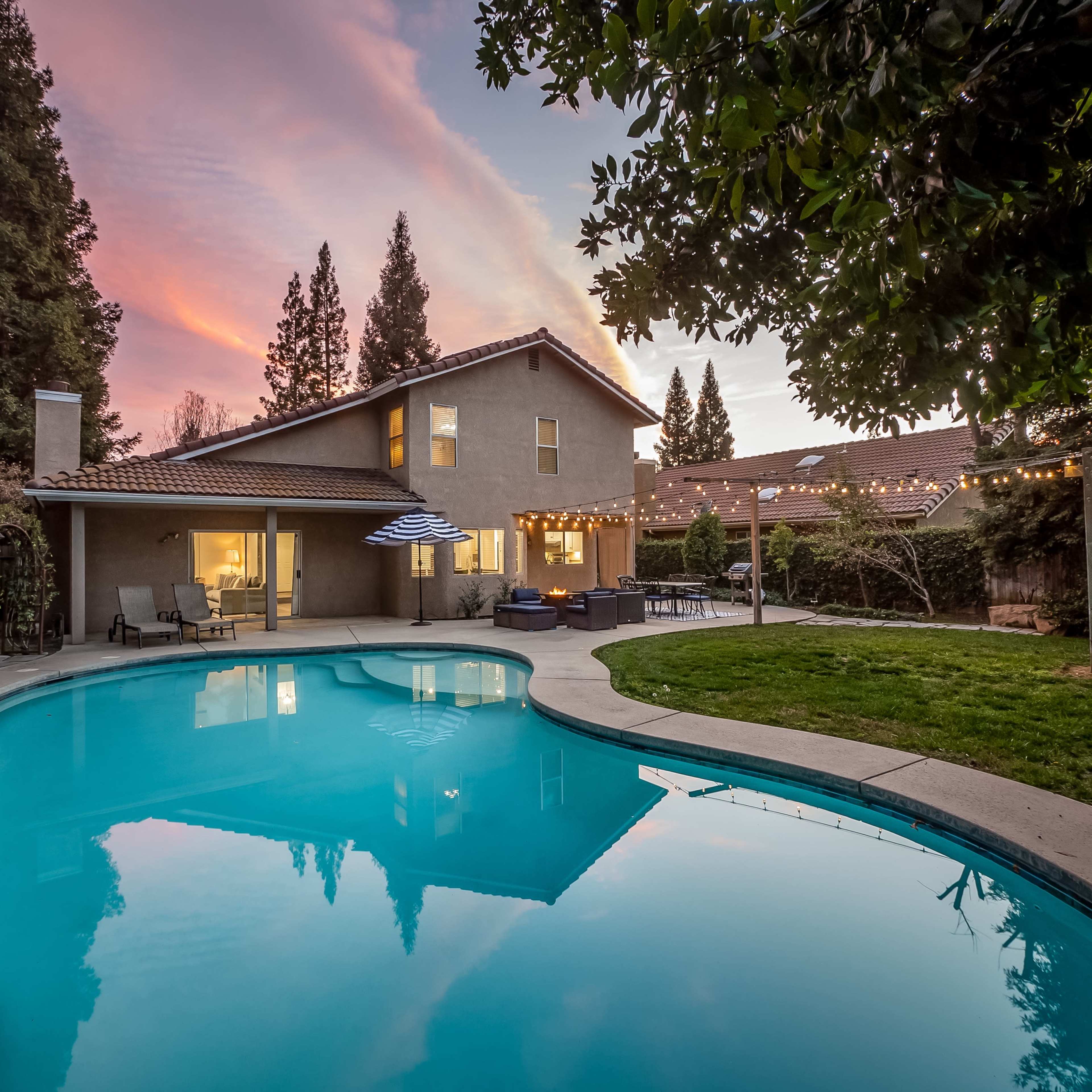 A house with a swimming pool is illuminated by evening lights and surrounded by trees and greenery.
