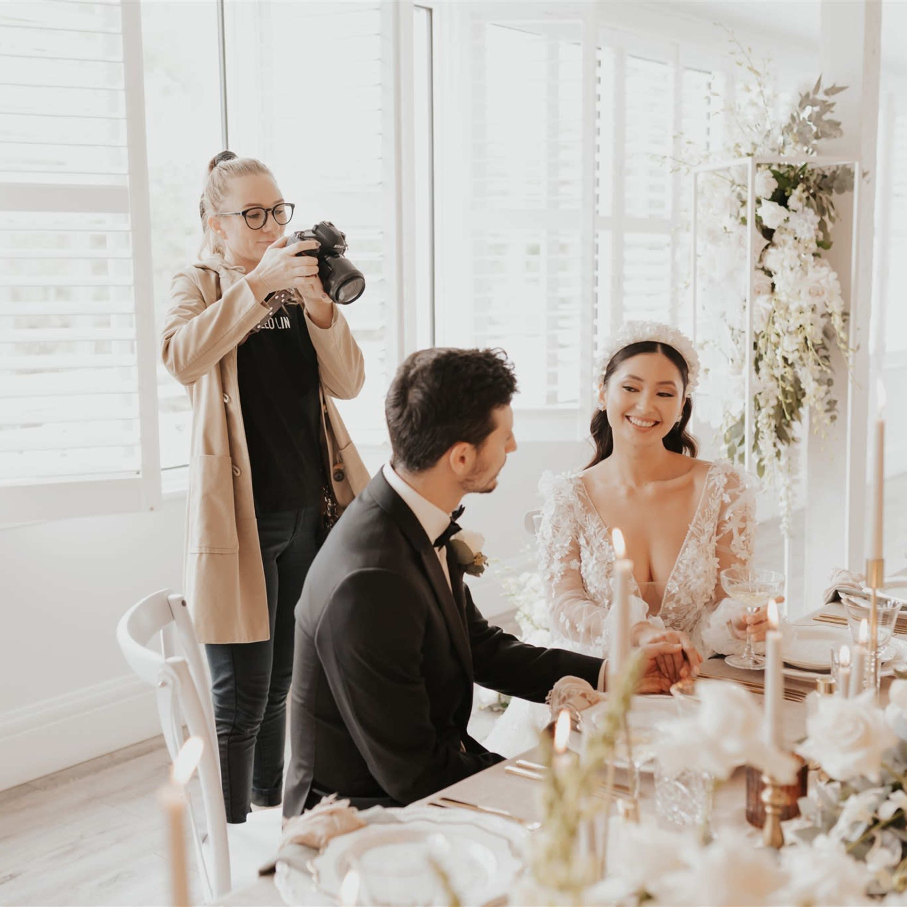 A couple is sitting at a beautifully set table decorated with flowers, while a photographer captures the moment in the background.