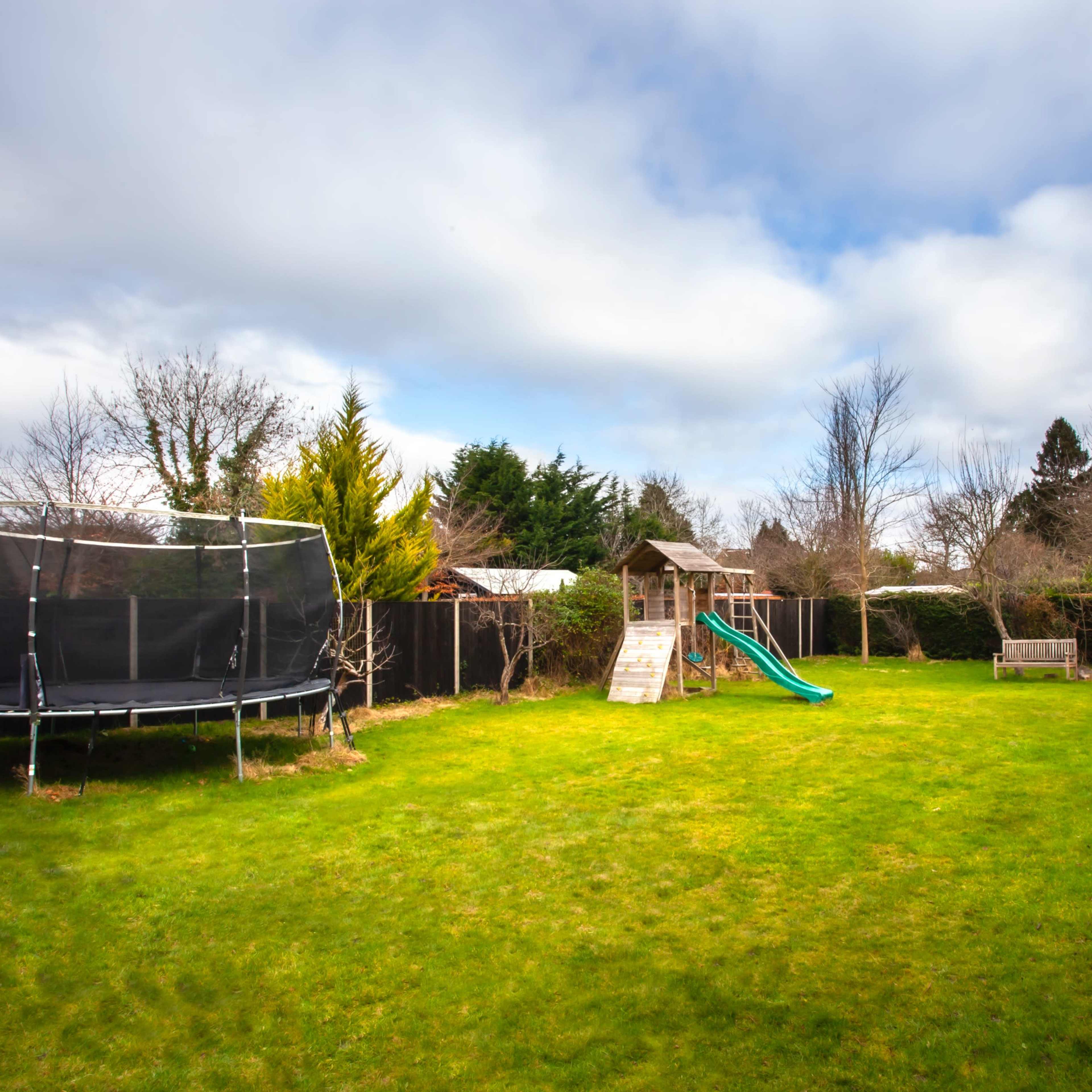 A spacious backyard with a trampoline, a playset with a slide, and a pathway bordered by shrubs.
