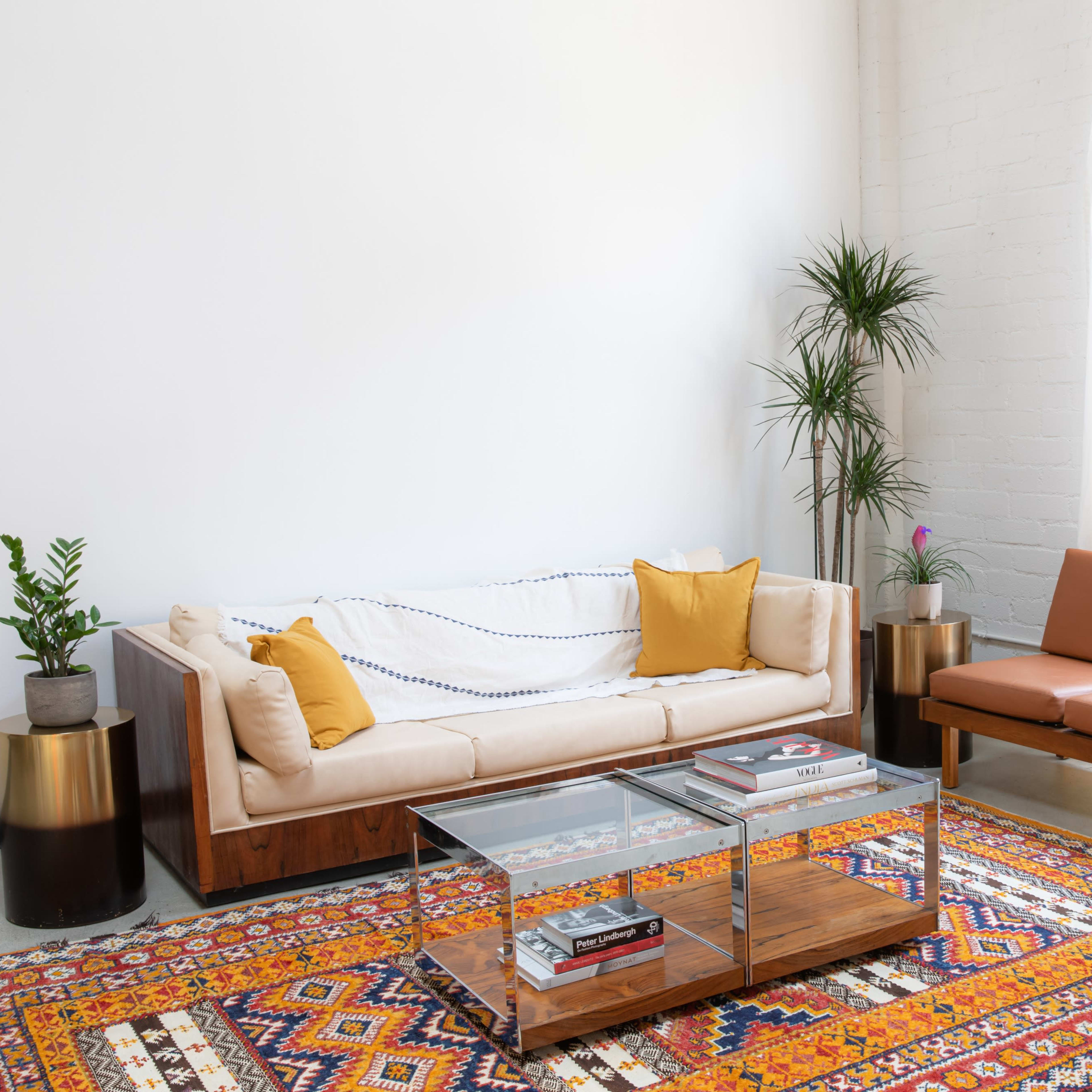A living room features a beige sofa with yellow cushions, a brown leather chair, a glass coffee table, and a colorful patterned rug, all against a backdrop of large windows and potted plants.