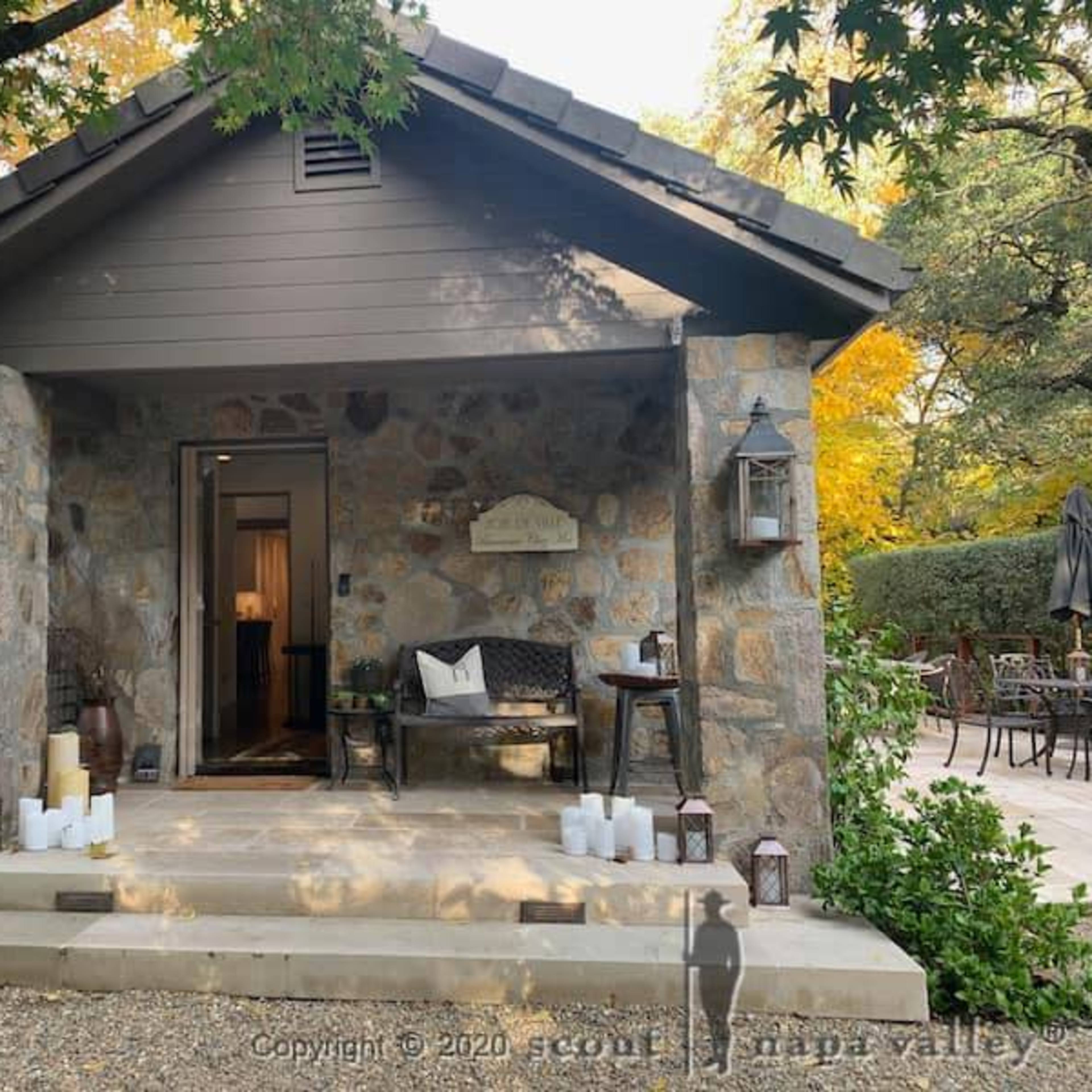 A stone cottage with a wooden porch, flanked by lanterns and white candles, is set amidst a landscaped garden.