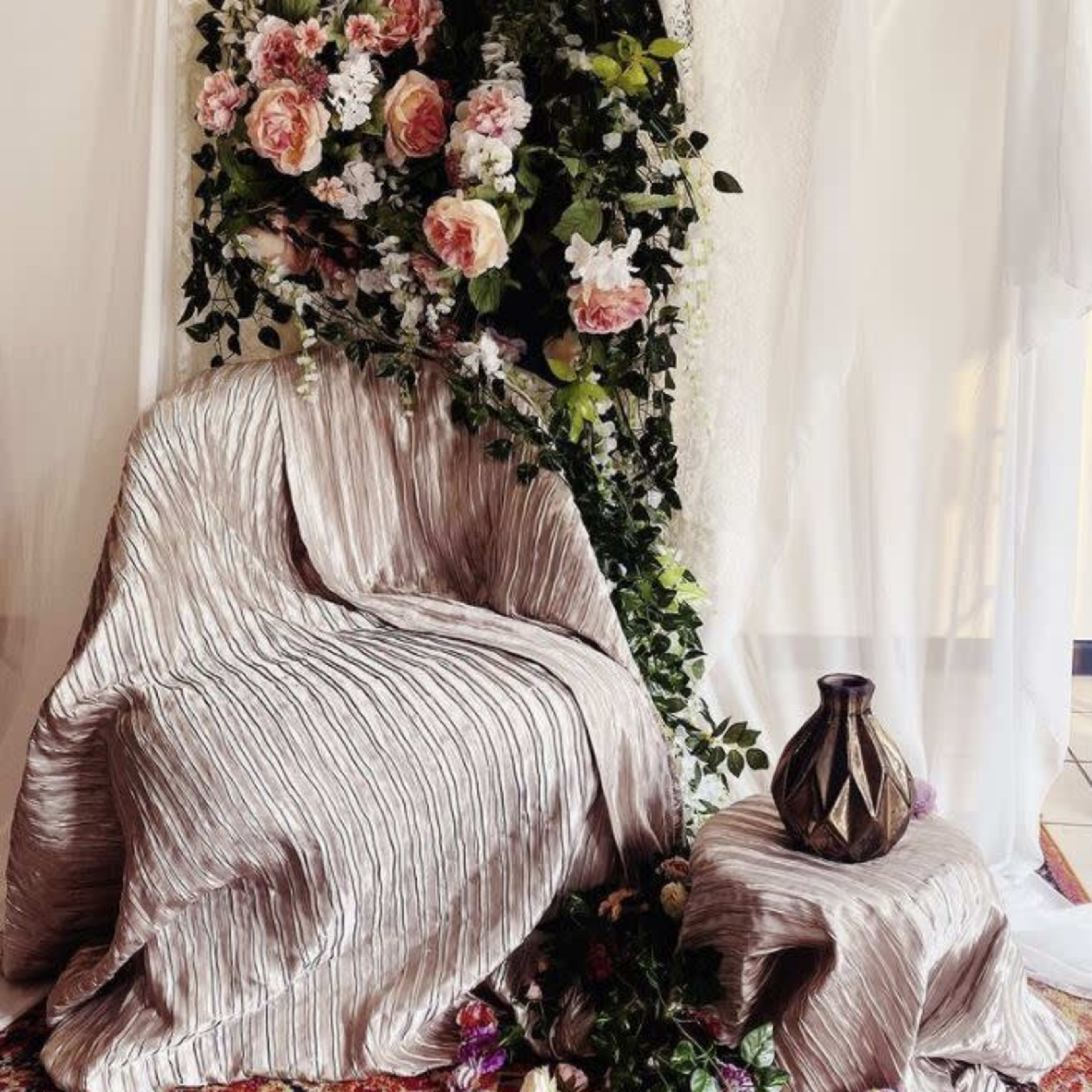 A draped chair and side table surrounded by floral decorations and a patterned rug.