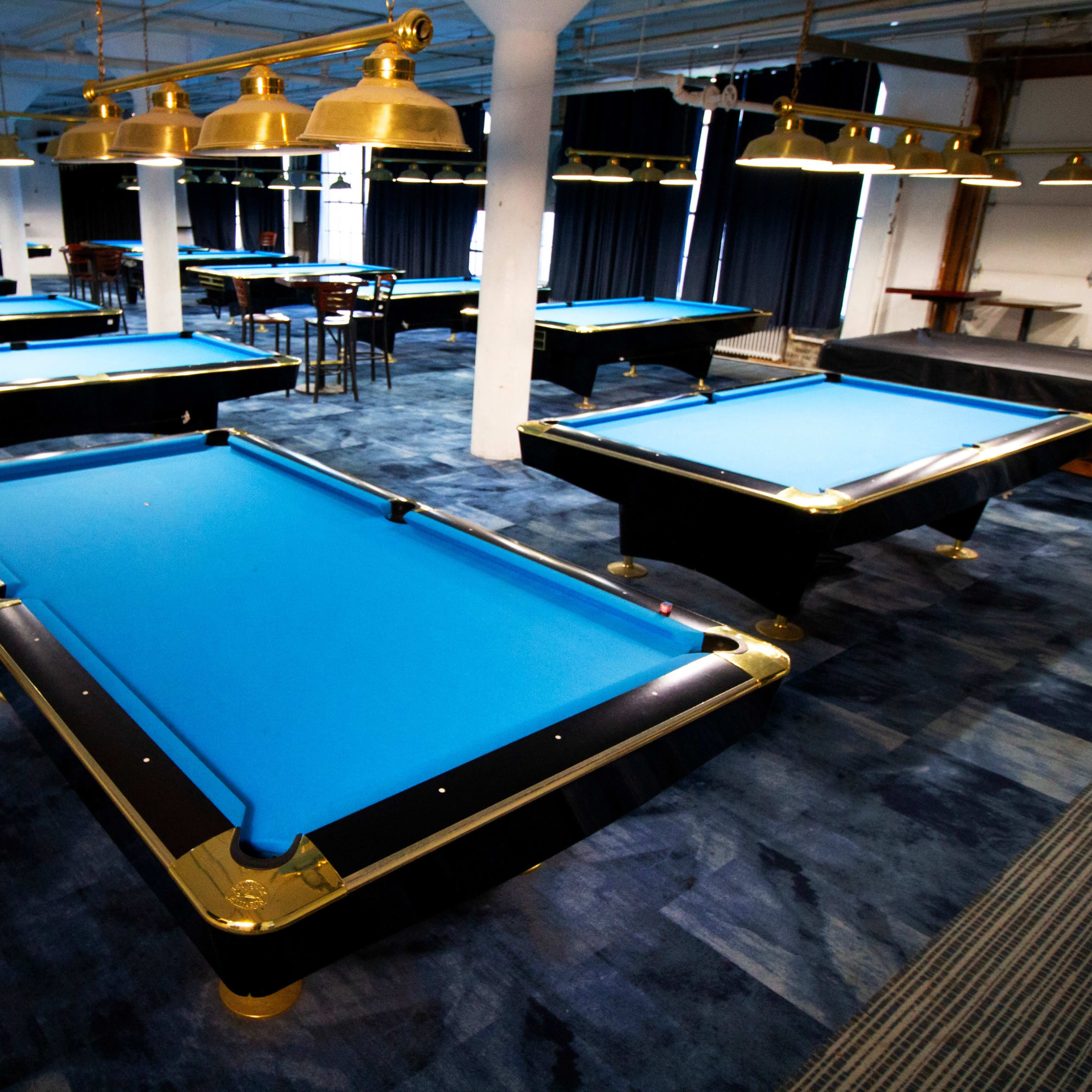 A spacious room filled with multiple pool tables, each covered with blue felt, arranged neatly on a dark blue carpet.
