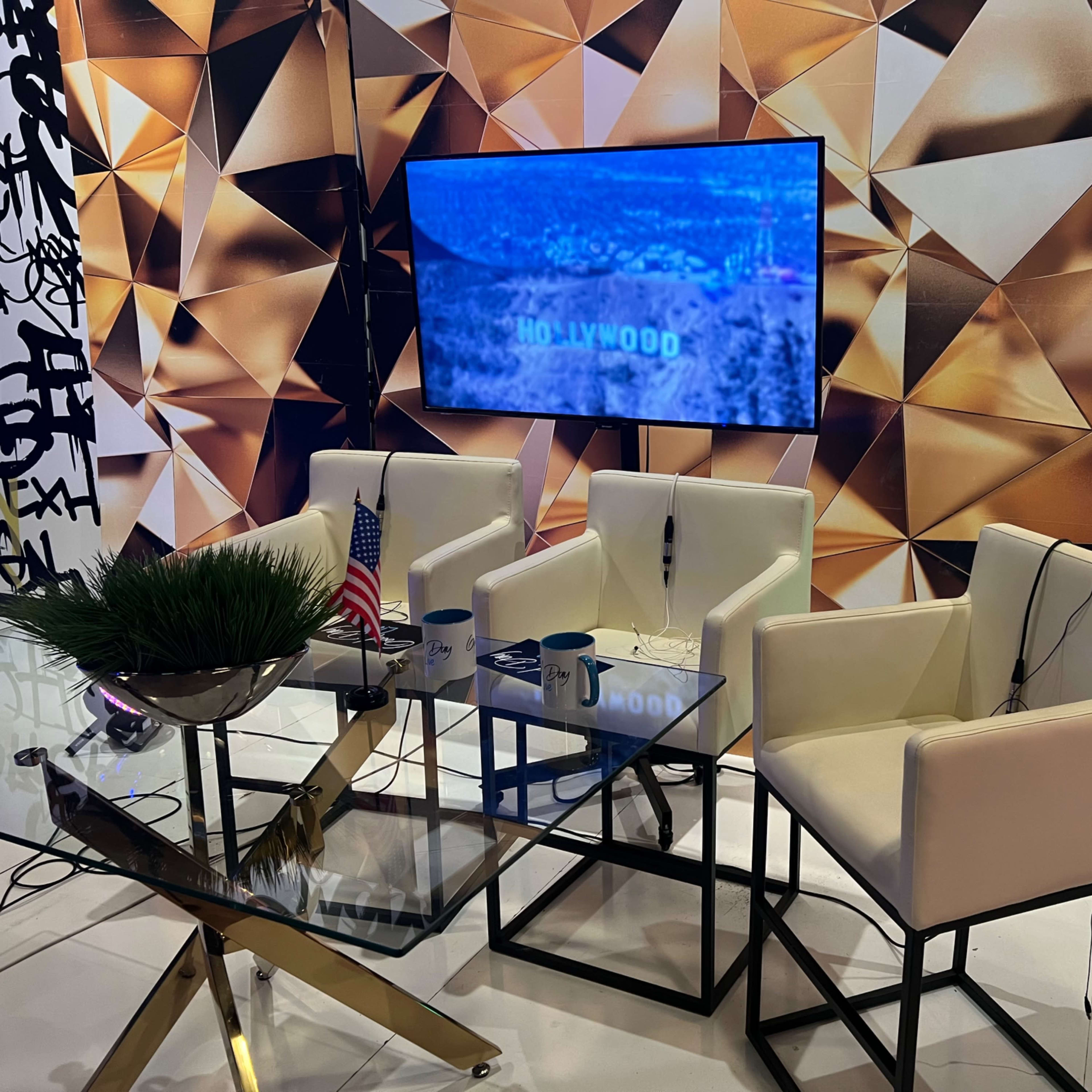 A modern interview setup featuring three white chairs, a glass table, an American flag, and a large screen displaying "HOLLYWOOD" against a geometric gold backdrop.