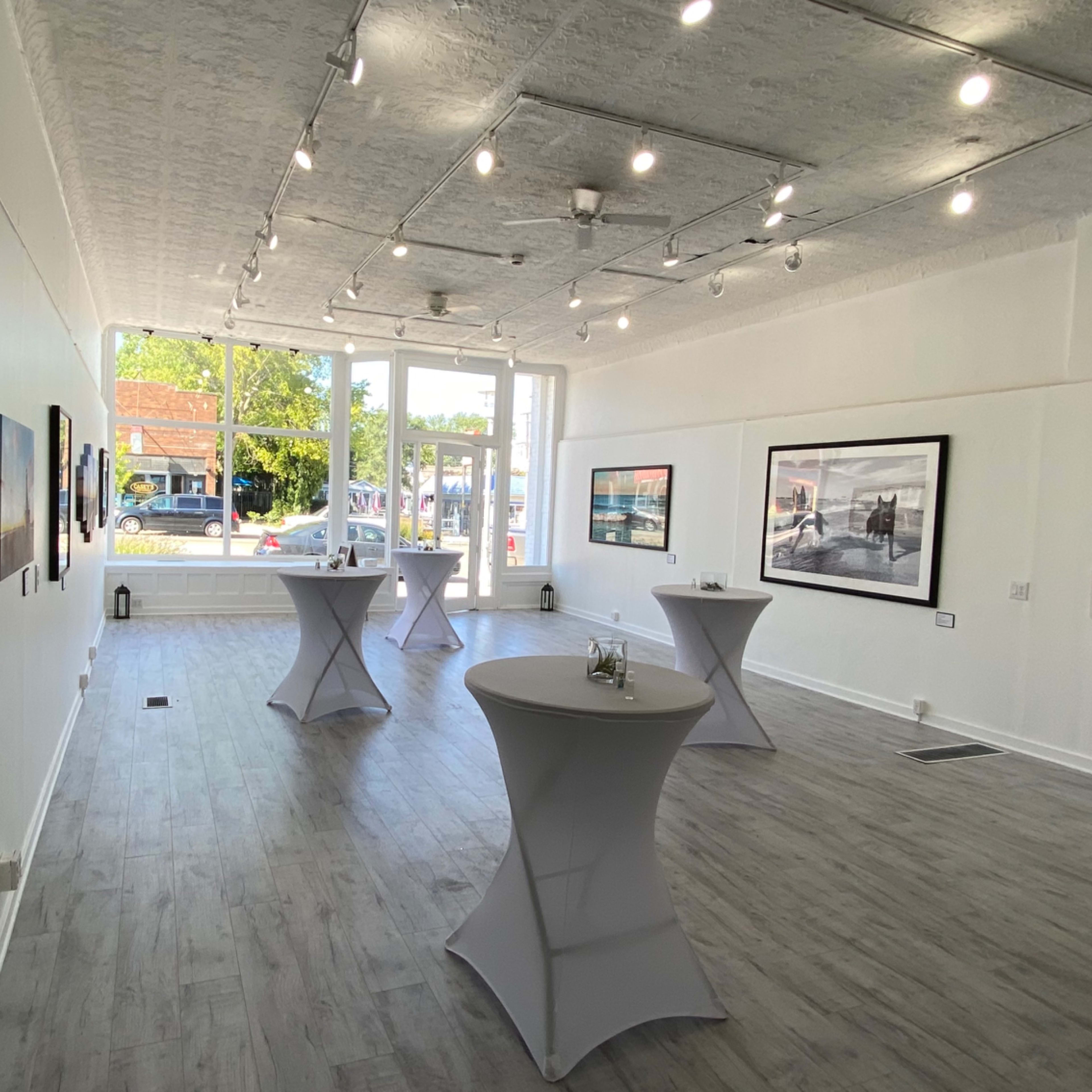 An art gallery with several framed artworks displayed on white walls, accompanied by tall tables for guests.