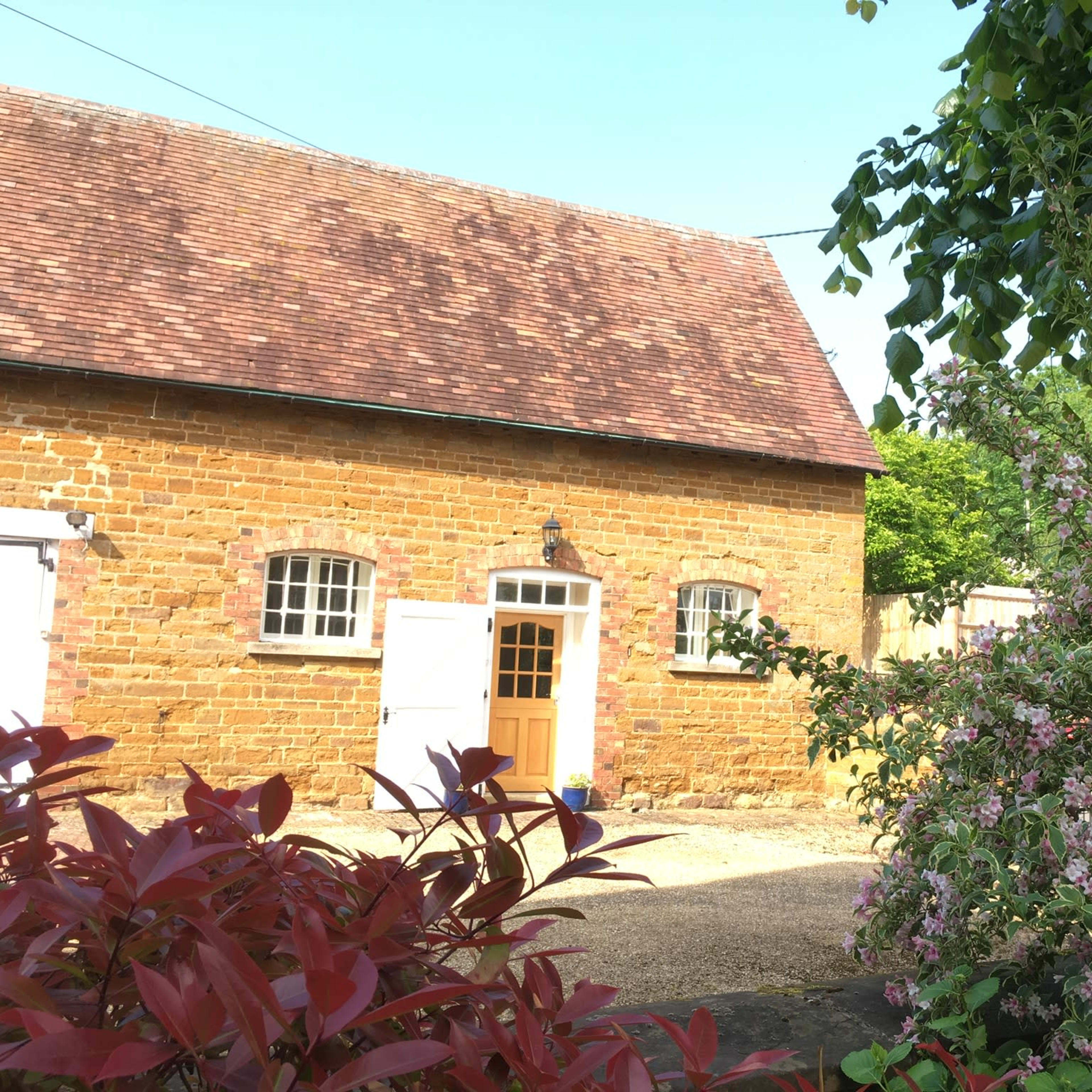 A stone cottage with a brown roof is surrounded by flowering plants and greenery.
