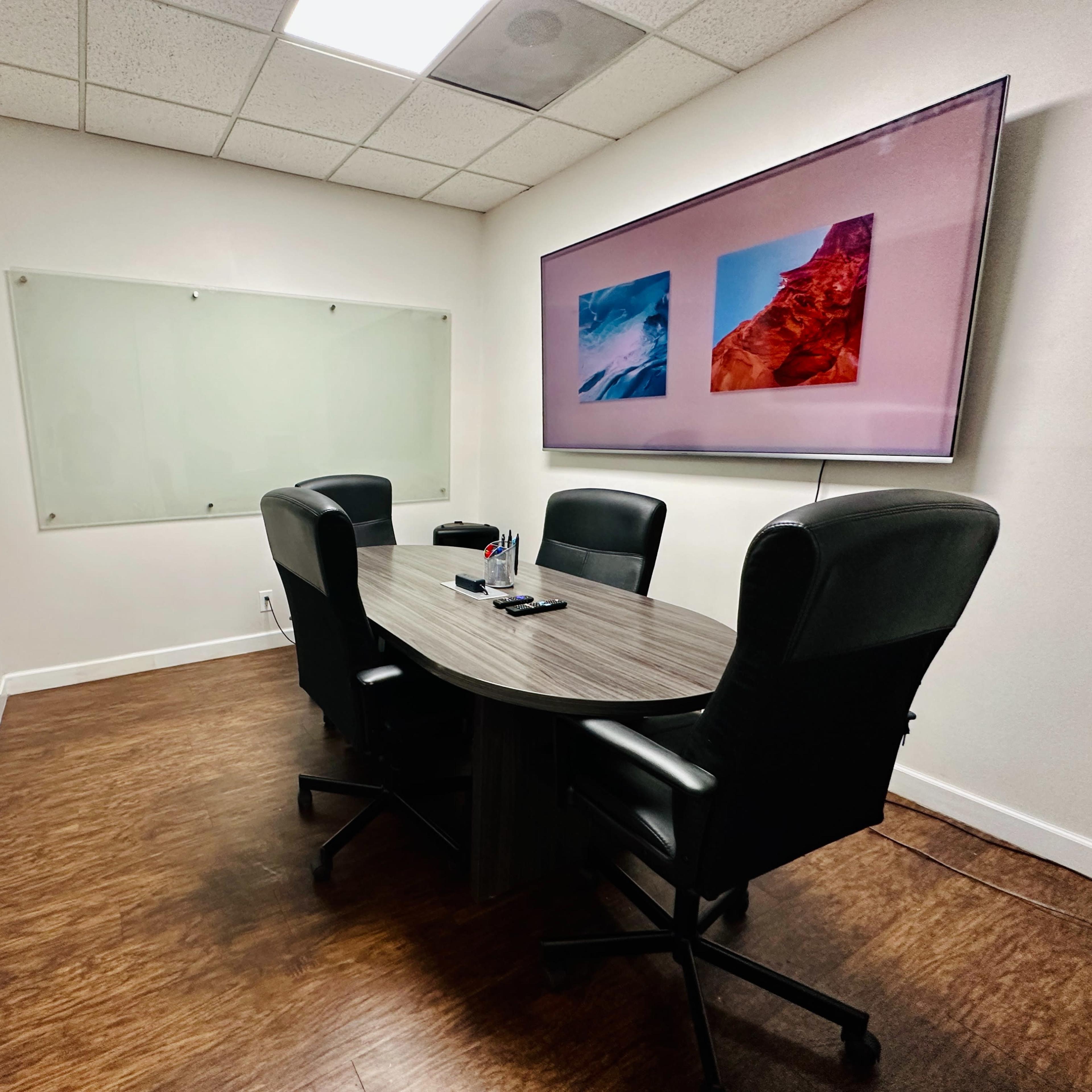 A small meeting room with a round table, four black chairs, a large screen displaying two images, and a whiteboard on one wall.