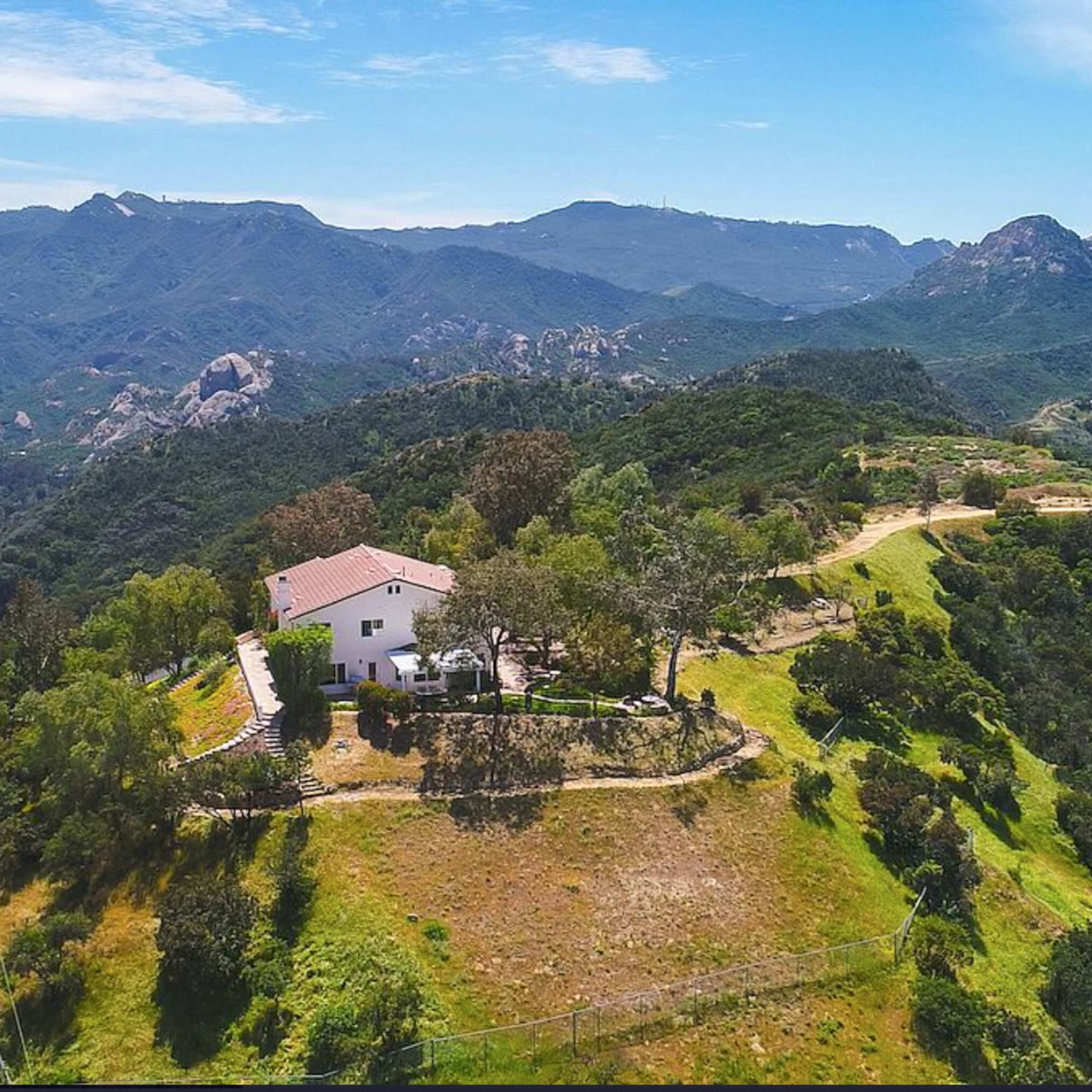 A single-story house is situated on a hillside, surrounded by trees and overlooking rolling mountains and valleys under a clear blue sky.