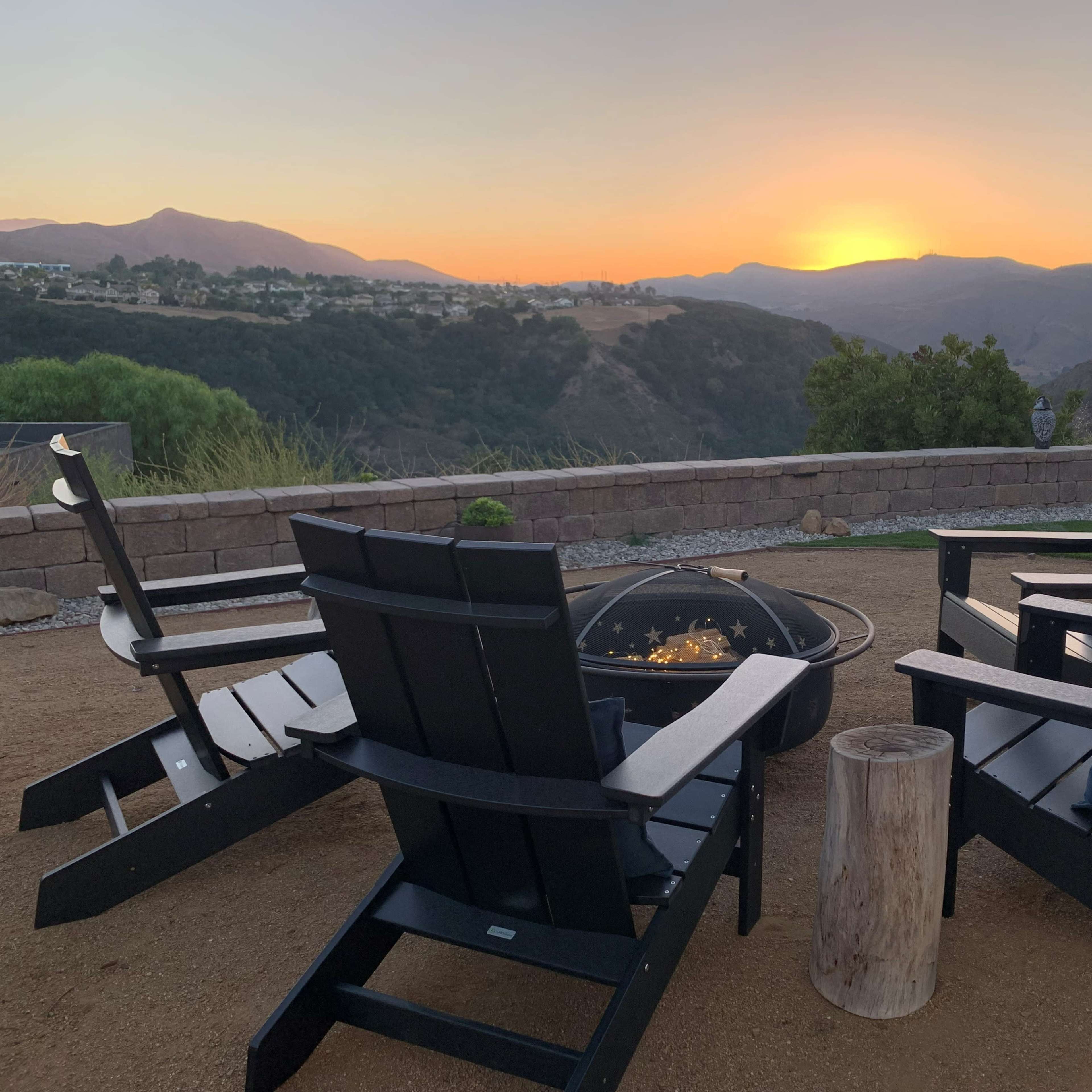 Four black Adirondack chairs surround a fire pit with a view of mountains and a sunset in the background.