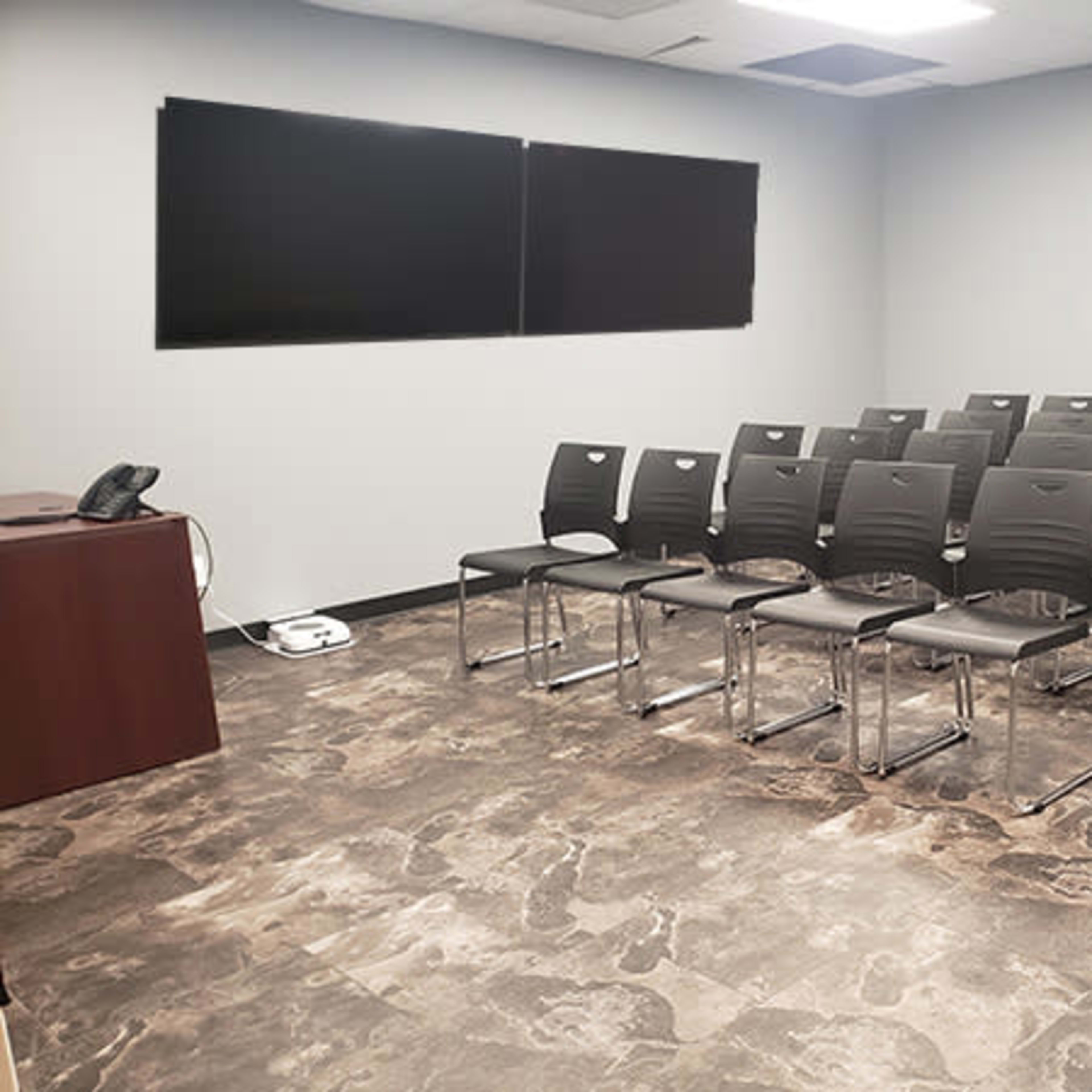 A conference room with a row of black chairs arranged in front of two large screens and a wooden table with a phone.