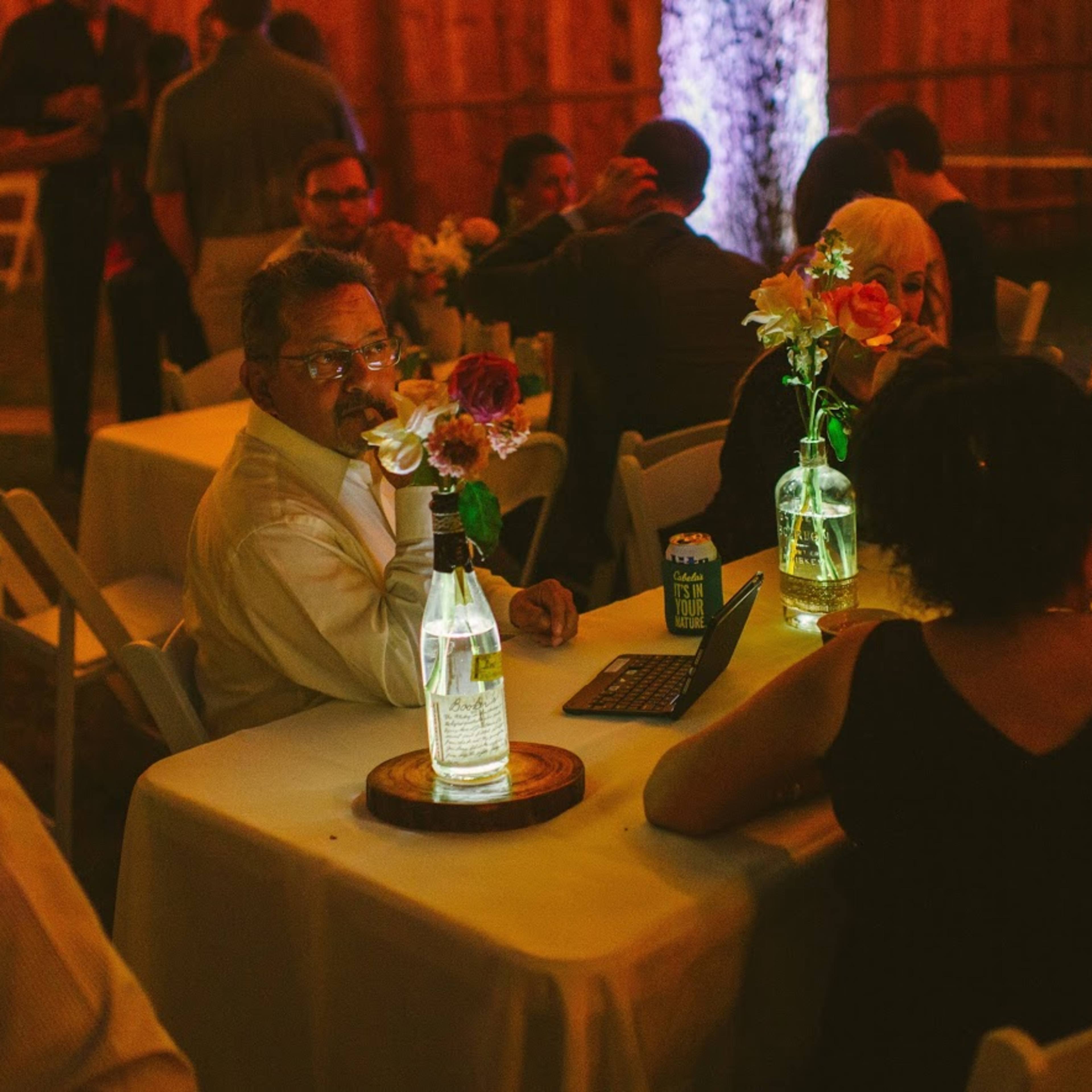 A group of people sits at a table decorated with flowers and illuminated water bottles in a dimly lit outdoor setting.