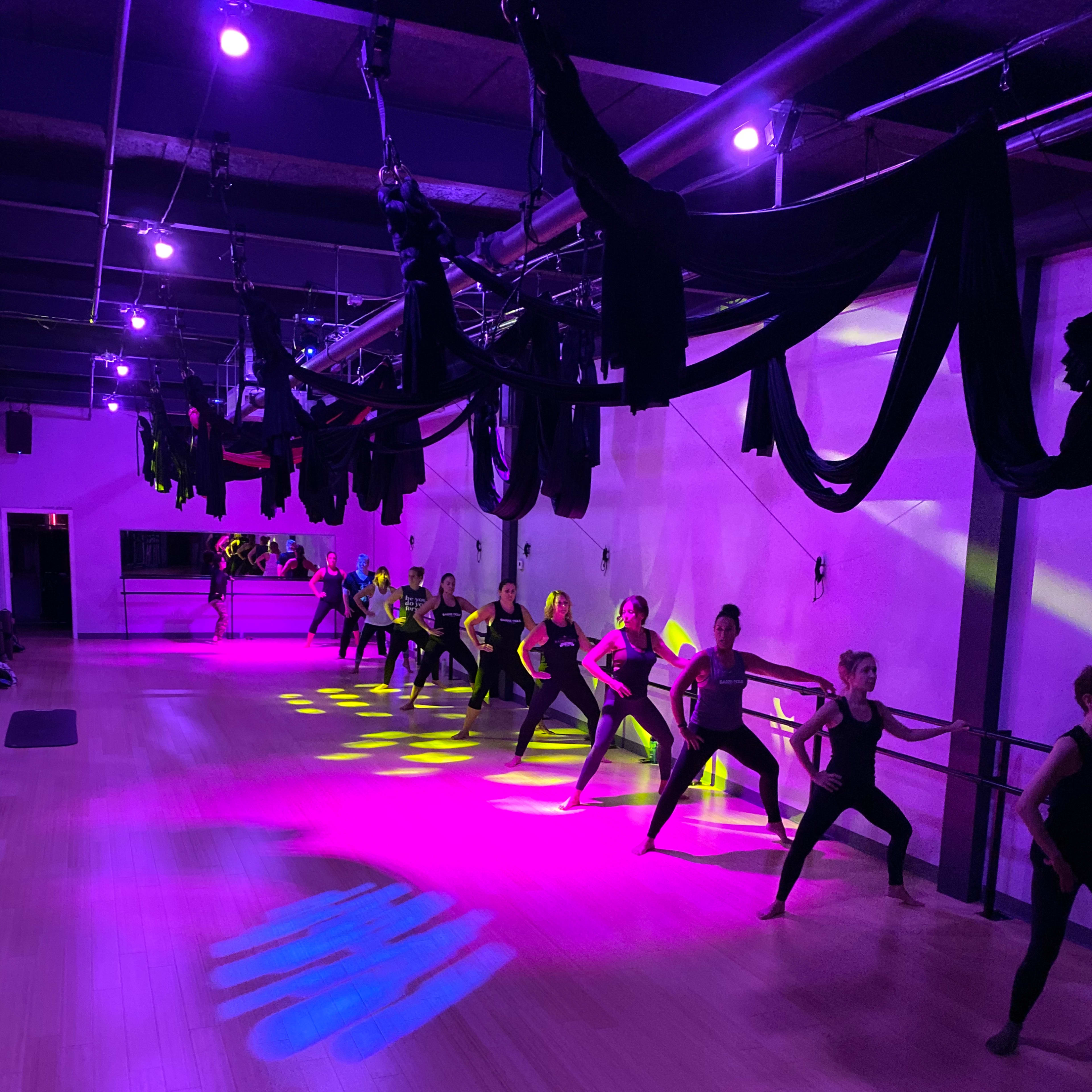 A dance studio features a group of dancers in formation under purple lights, with colorful shadows displayed on the floor.