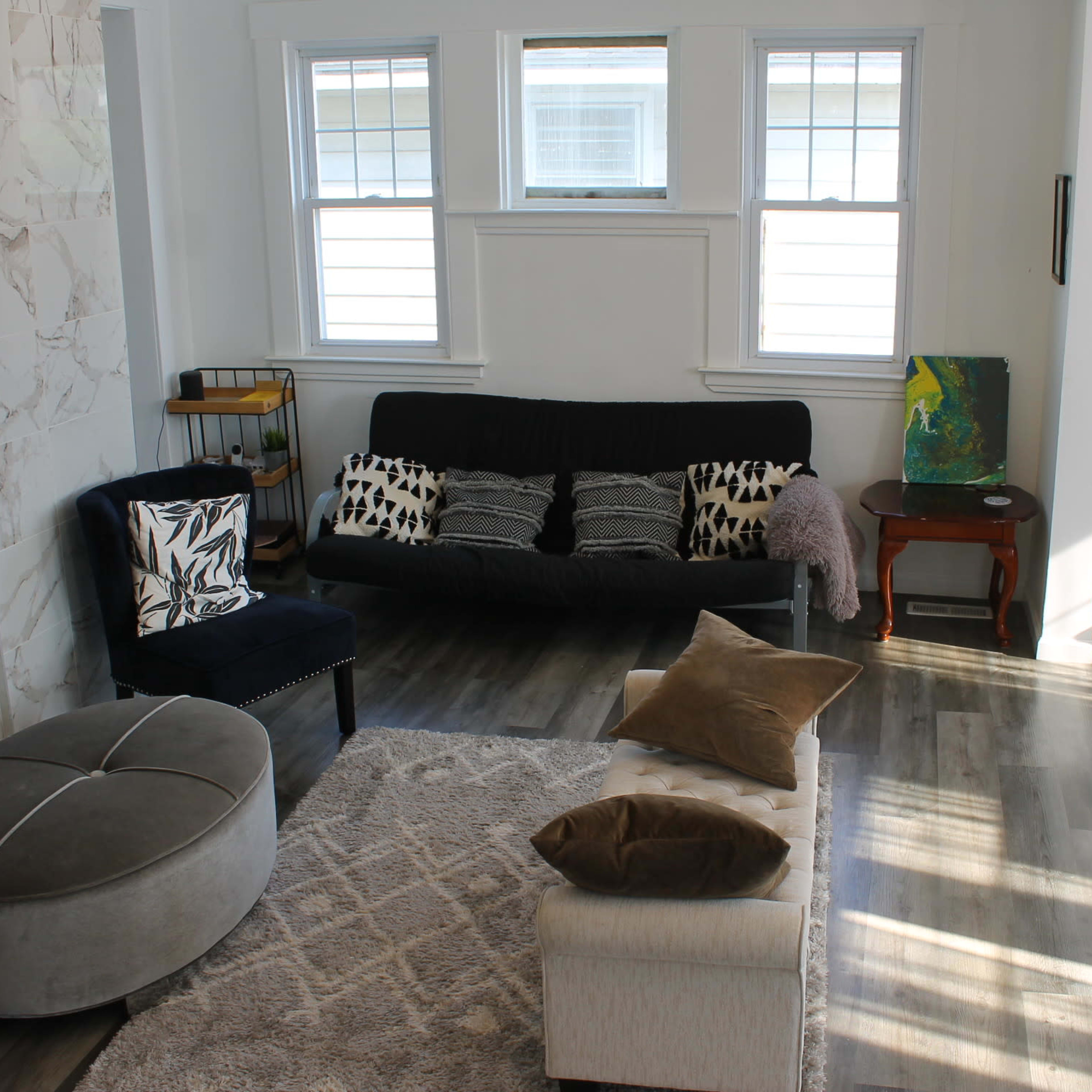 A living room with a sofa, armchairs, a round ottoman, and a coffee table, all arranged on a light-colored rug with wood flooring.