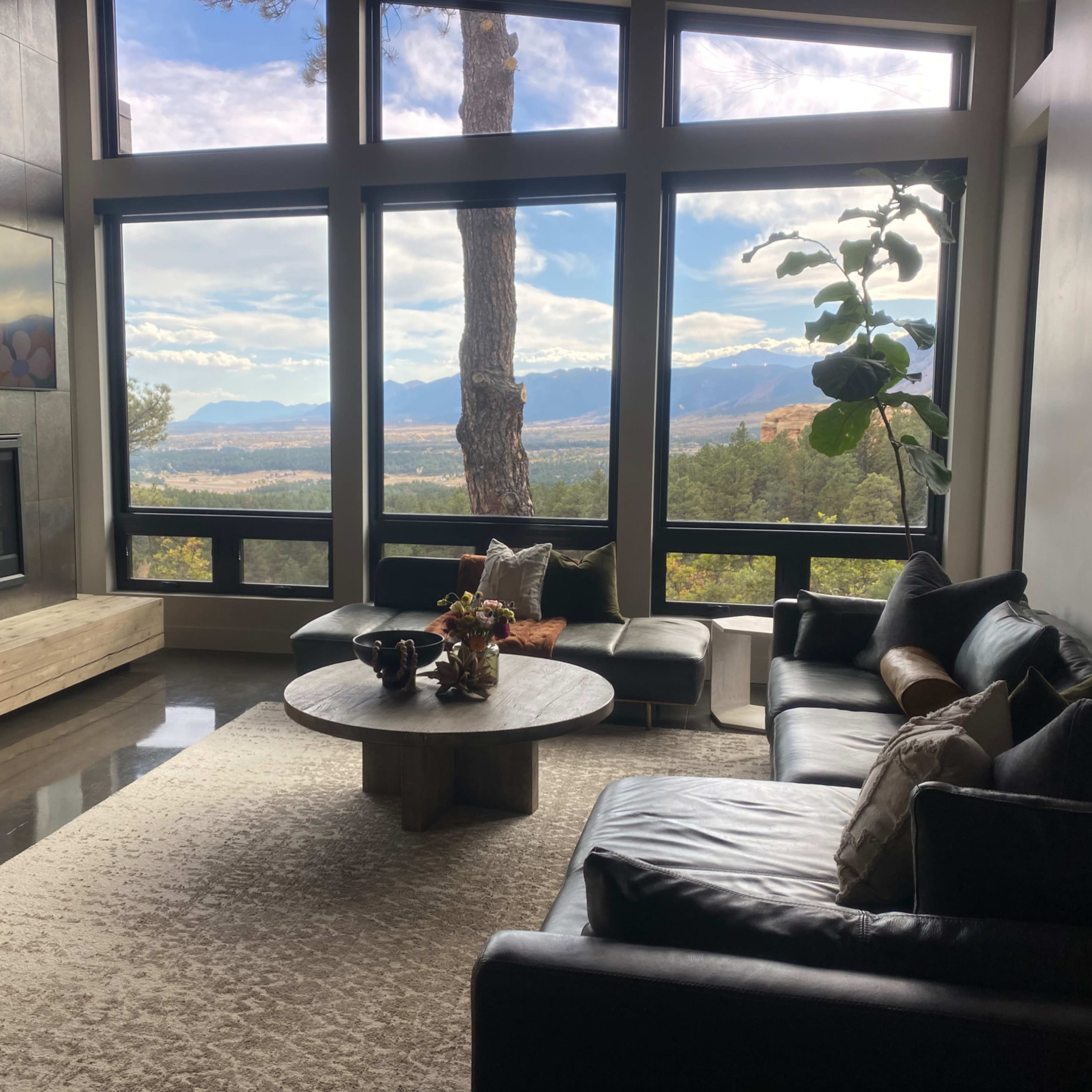 A modern living room features a large window with views of mountains and trees, a circular coffee table, and a black leather sofa arrangement.