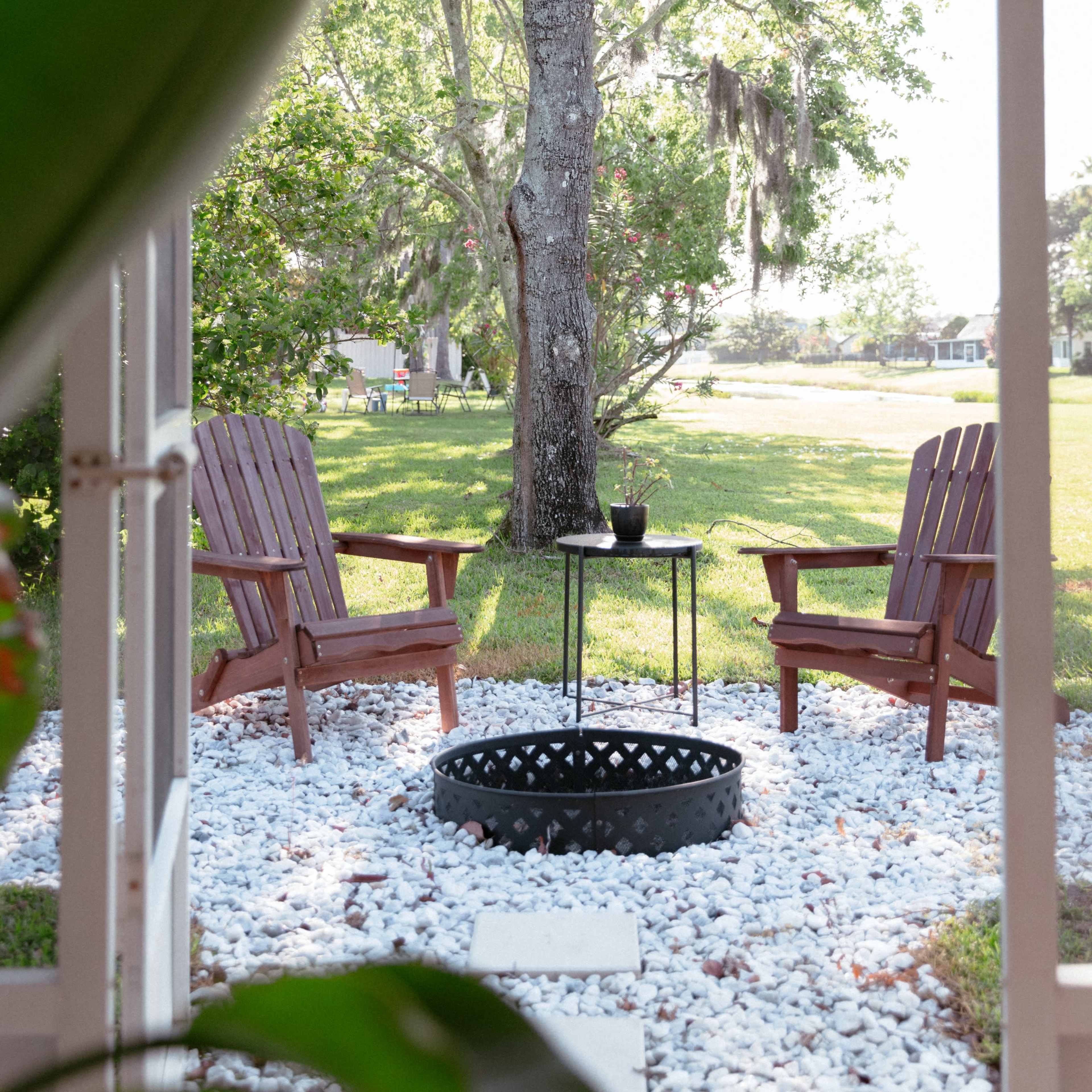 Two wooden chairs are positioned on a gravel area under a tree, with a small table in between them.